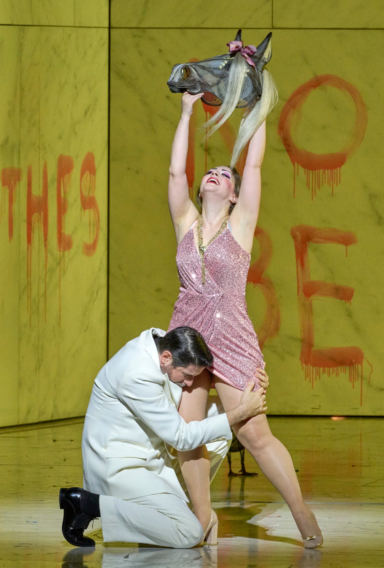 Eine Frau in glitzerndem Kleid hebt triumphierend eine Pferdemaske über den Kopf, während ein Mann sich vor ihr niederbeugt; im Hintergrund rote Schrift an gelber Wand.