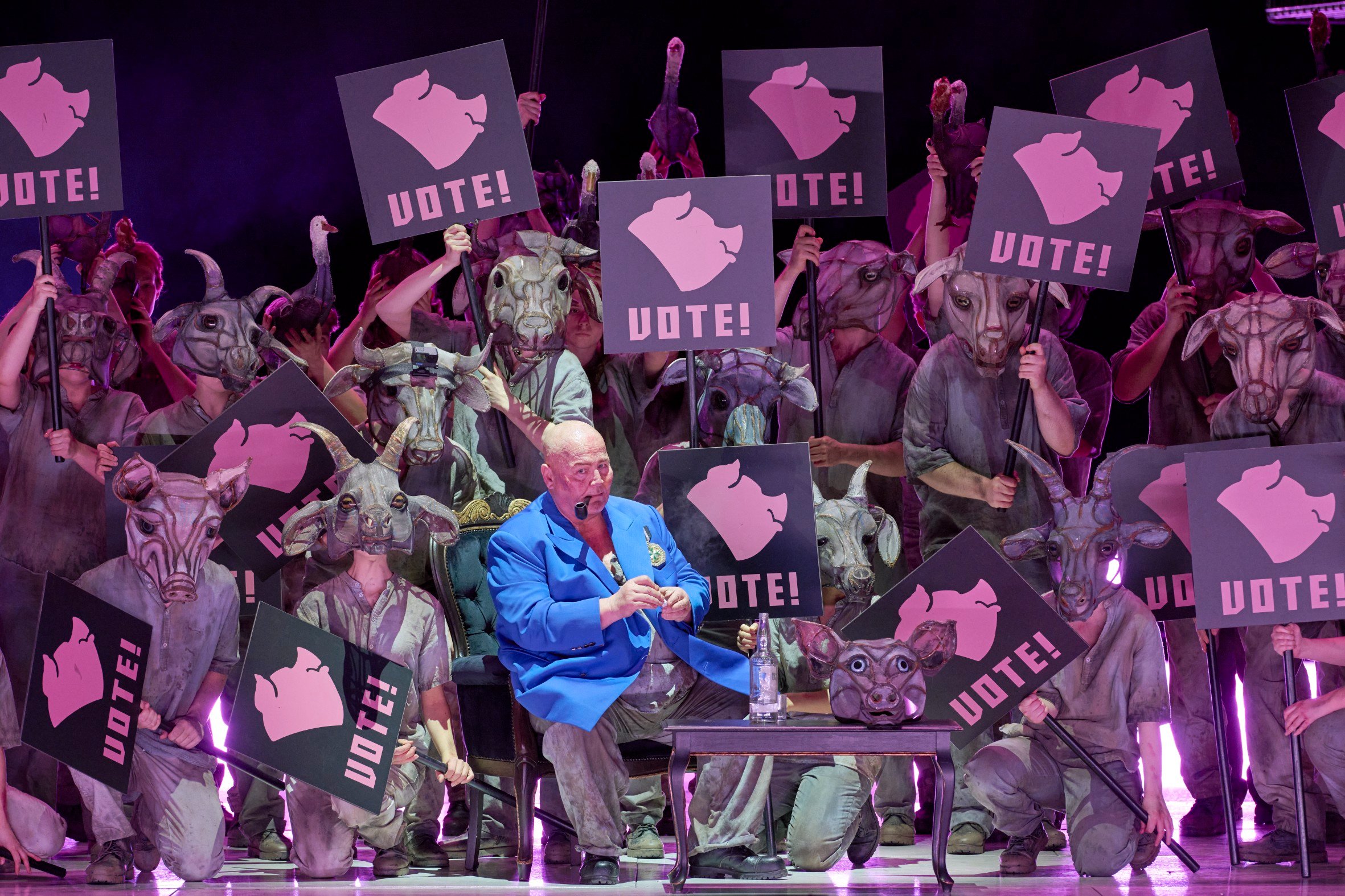 Ein Mann im blauen Sakko sitzt vor einer Gruppe maskierter Figuren, die rosa Schilder mit der Aufschrift „VOTE!“ und einem Schweinekopf-Symbol hochhalten.