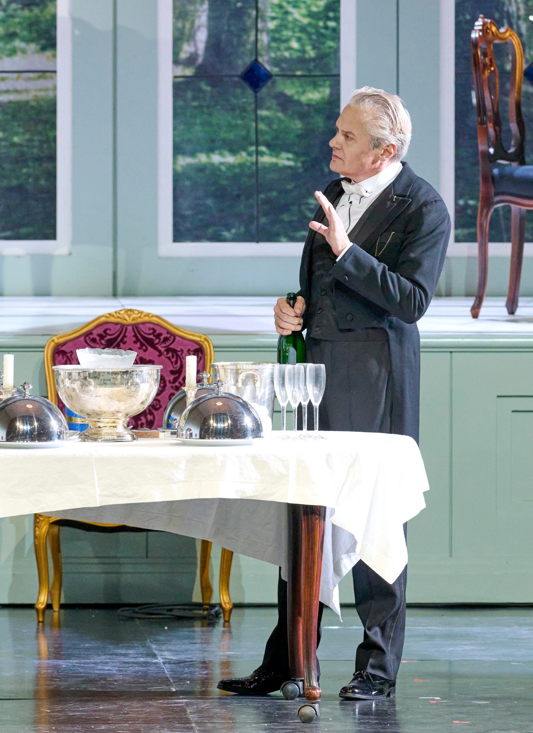 Ein elegant gekleideter Mann steht hinter einem weiß gedeckten Tisch mit Sektgläsern und Silberhauben, eine geöffnete Champagnerflasche in der Hand.