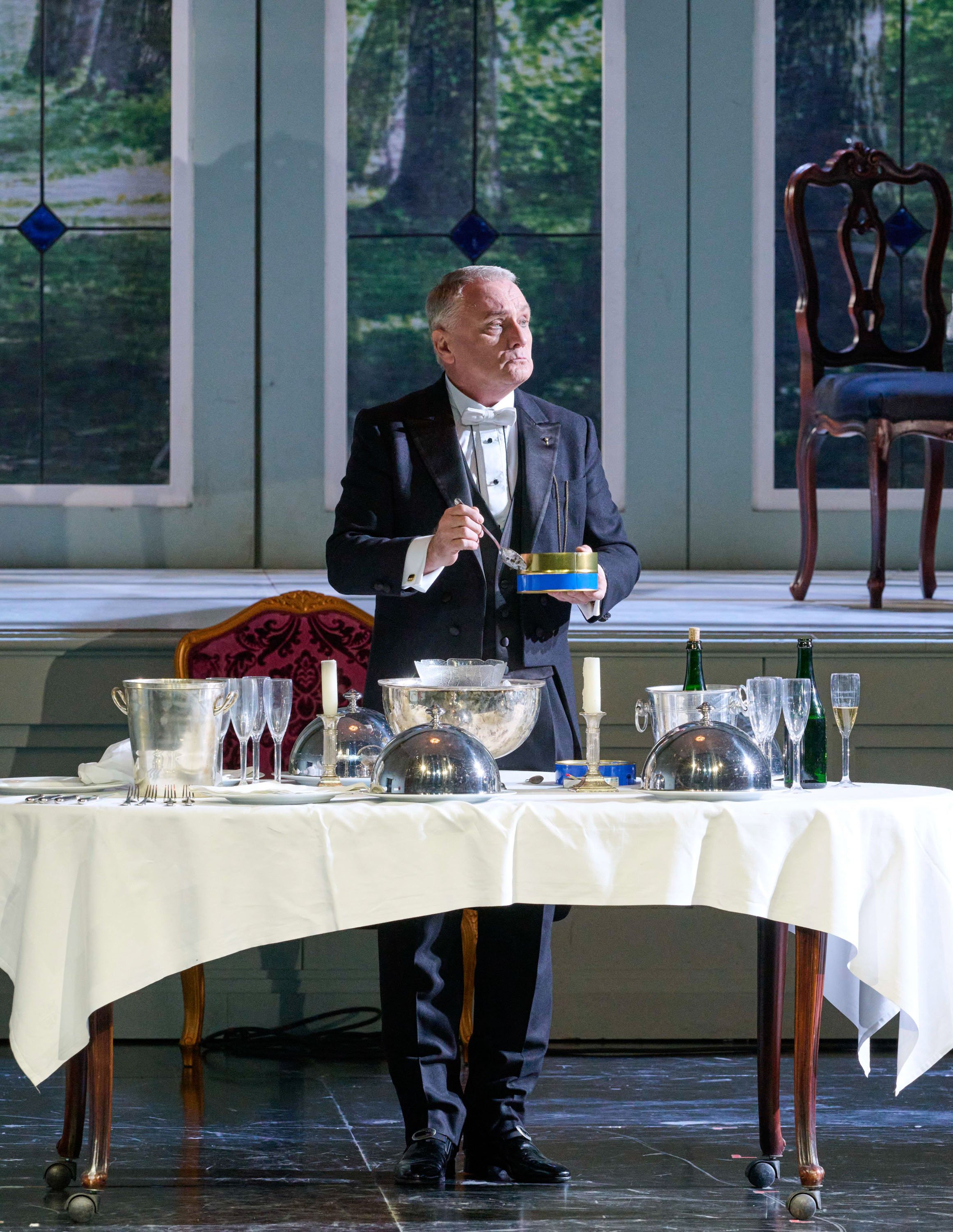 Ein älterer Mann im Smoking steht hinter einem elegant gedeckten Tisch mit Champagner, Gläsern und silbernen Servierglocken, während Licht durch Fenster im Hintergrund fällt.