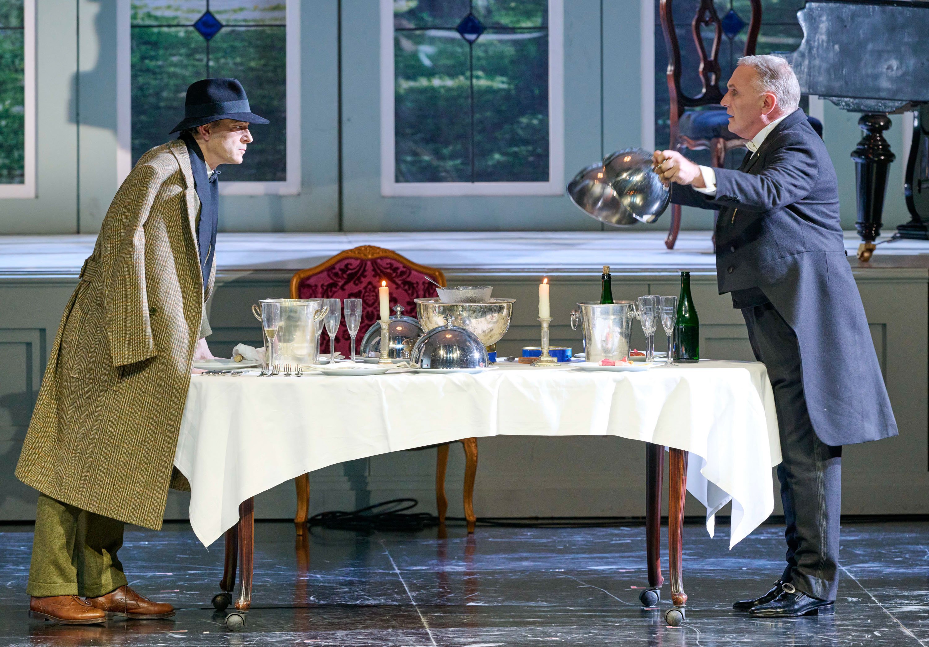 Zwei Männer stehen sich an einem reich gedeckten Tisch gegenüber; der eine hebt eine Servierglocke, während der andere in Mantel und Hut gespannt zusieht.