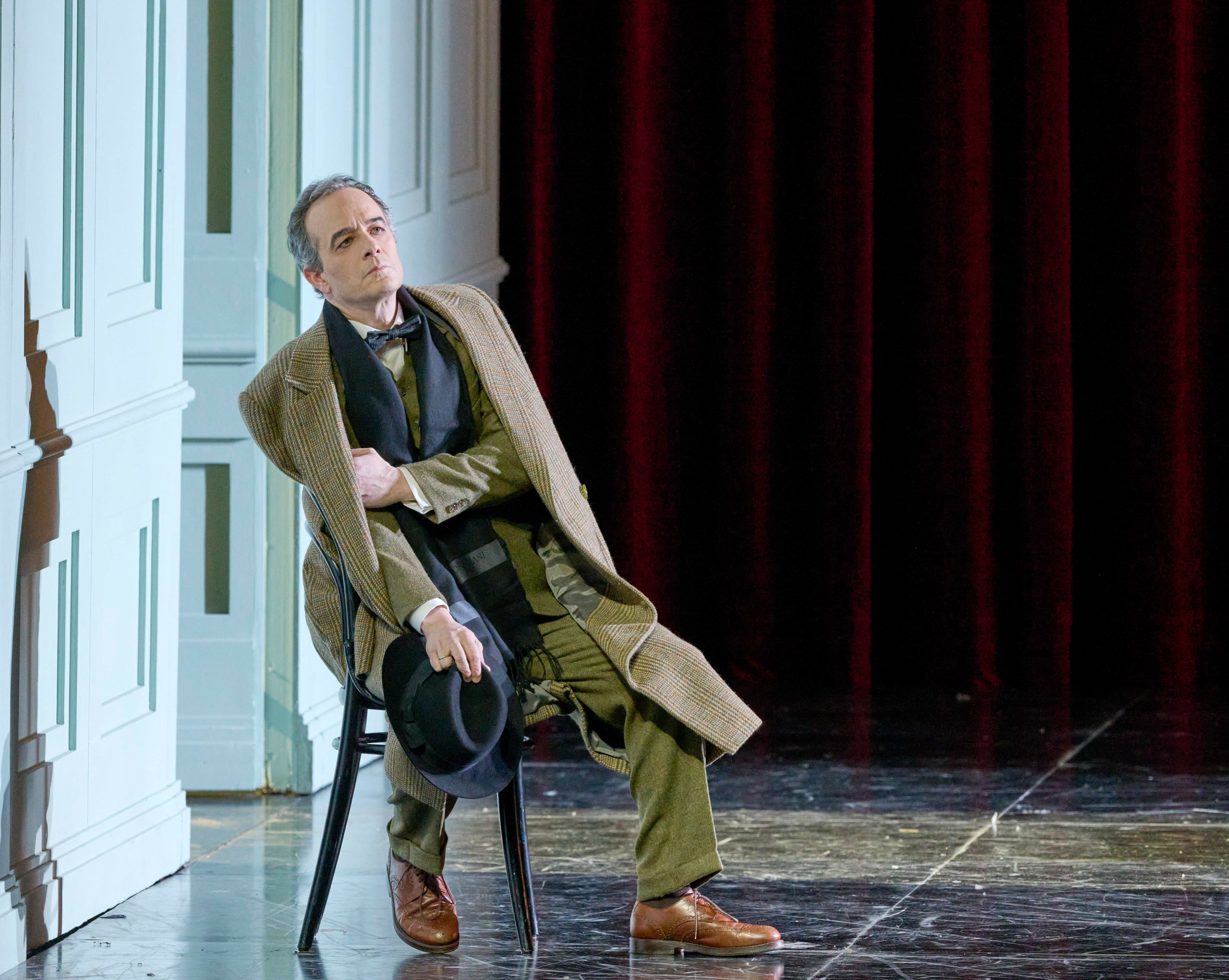 Ein Mann in langem Mantel und Anzug sitzt erschöpft auf einem Stuhl, hält einen Hut in der Hand und lehnt sich an die Wand eines hellen Bühnenraums mit rotem Vorhang.