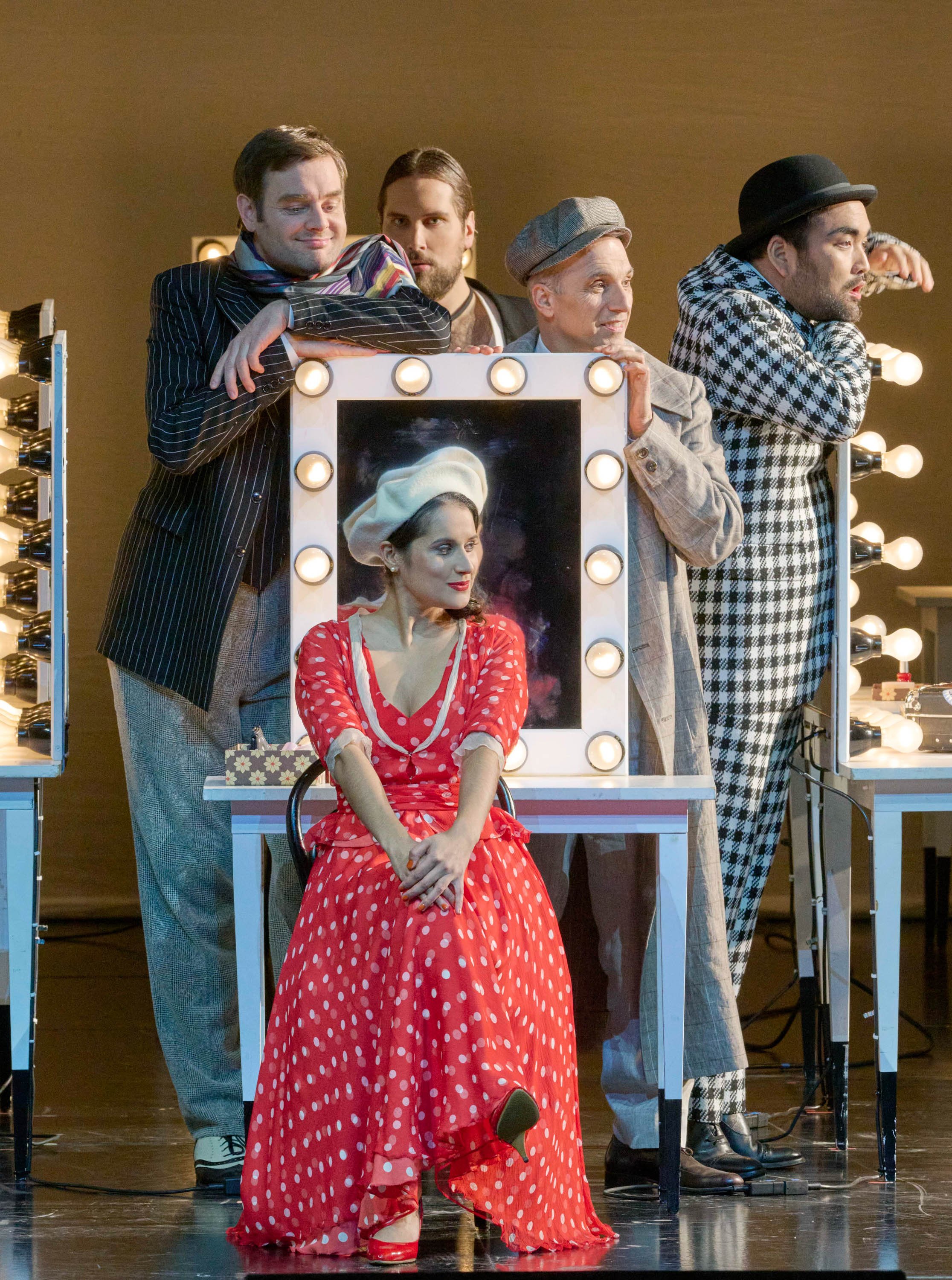 Eine Frau in rotem Kleid und Barett sitzt vor einem beleuchteten Schminktisch, während vier Männer in verschieden gemusterter Kleidung sich um sie gruppieren.