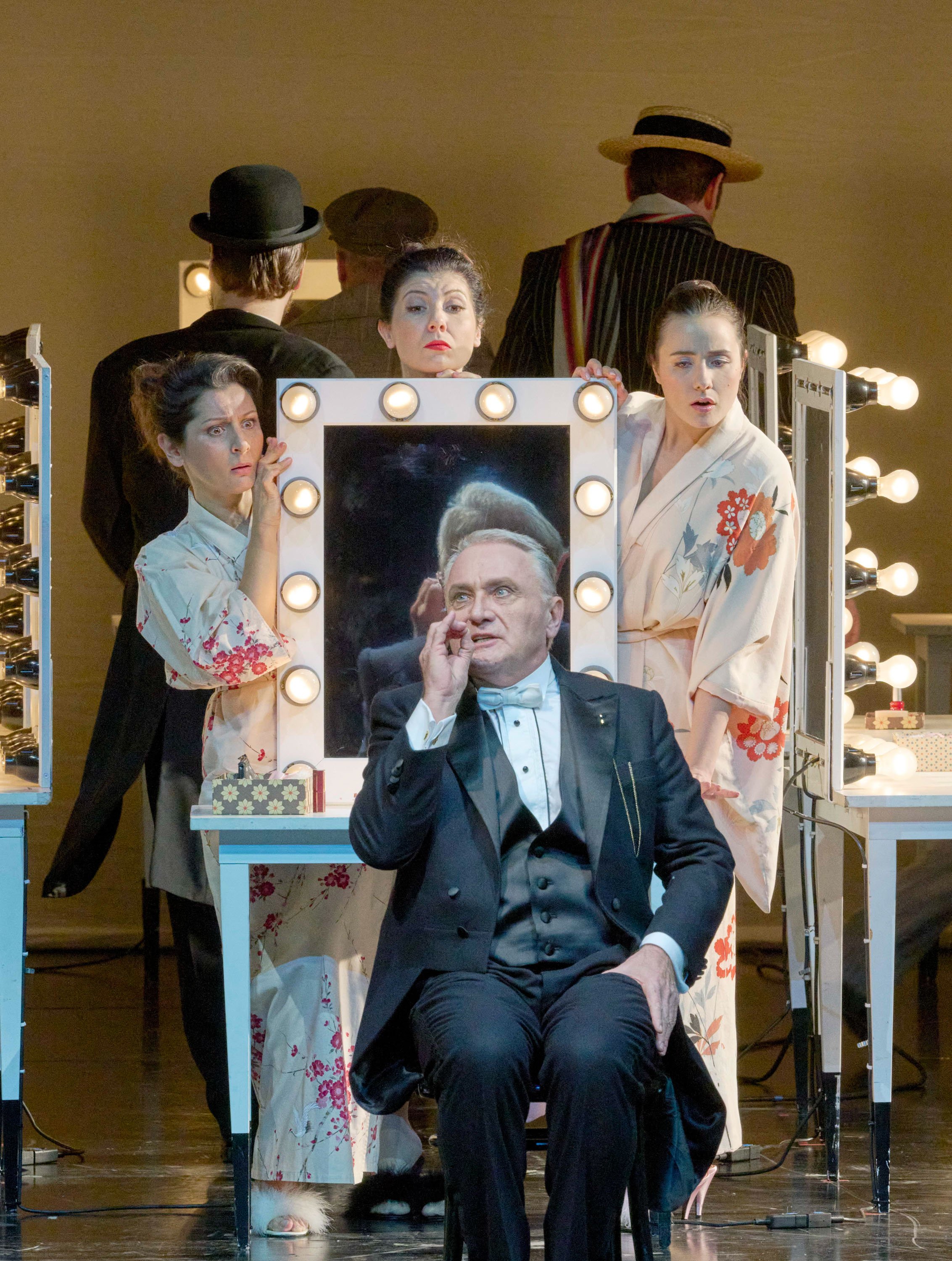 Ein Mann im Smoking sitzt selbstzufrieden vor einem beleuchteten Schminkspiegel, umgeben von drei Frauen in weißen Kimonos mit Blumenmuster.