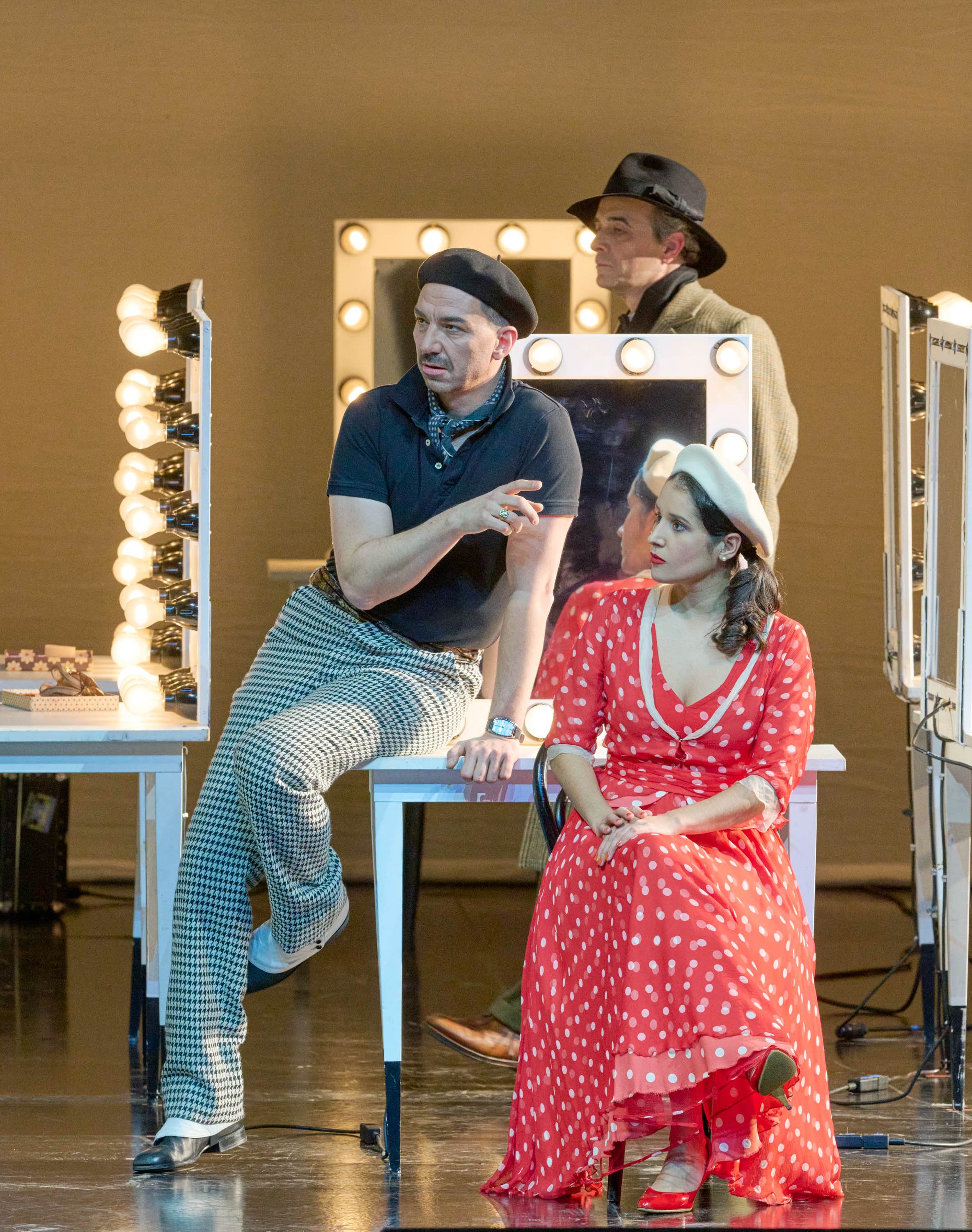 Eine Frau in rotem Kleid und Barett sitzt vor einem beleuchteten Schminktisch, während ein Mann mit schwarzem Shirt und Mütze neben ihr sitzt.