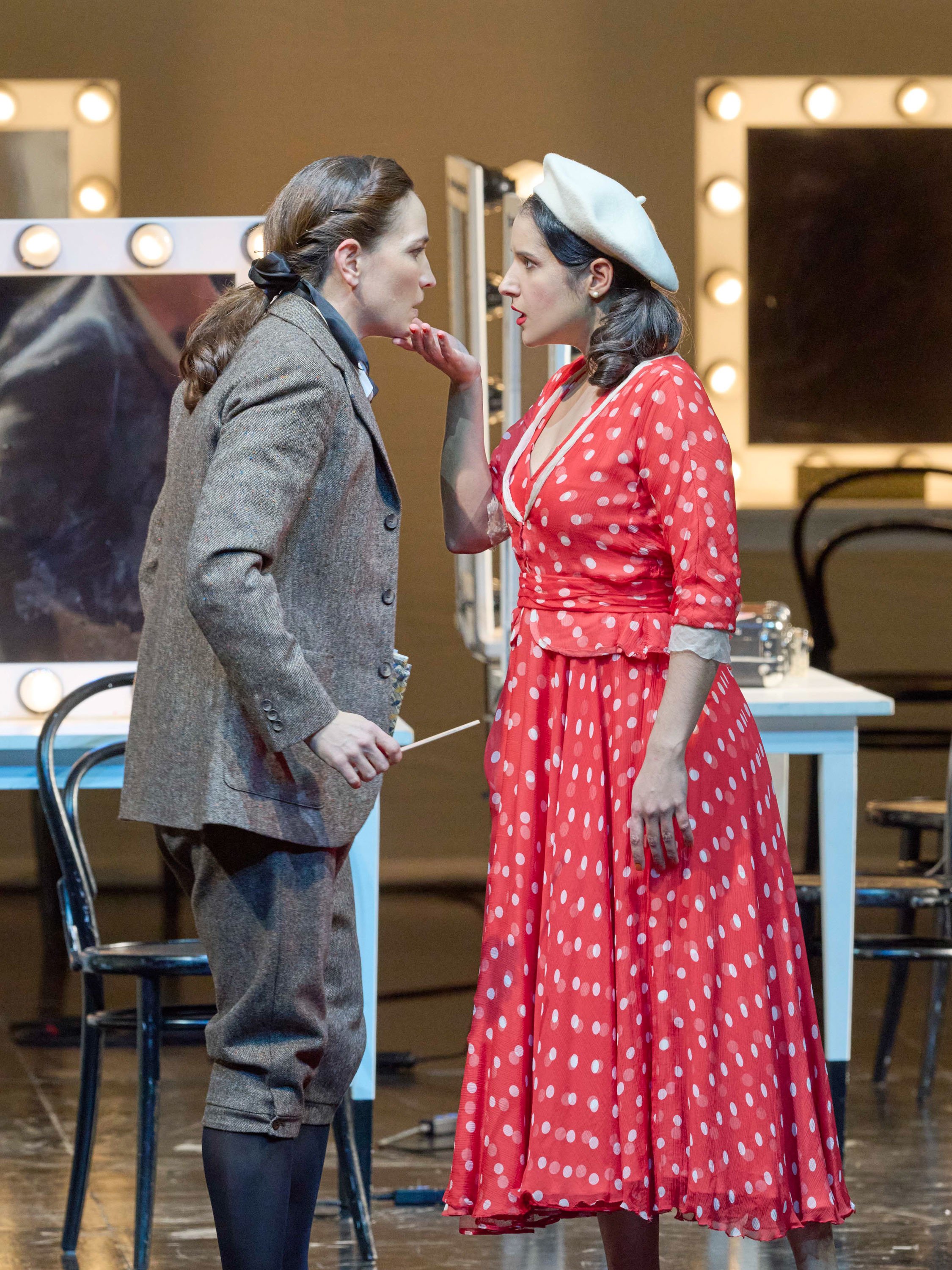 Zwei Frauen stehen sich vor einem beleuchteten Schminktisch gegenüber; die eine hebt sanft das Kinn der anderen