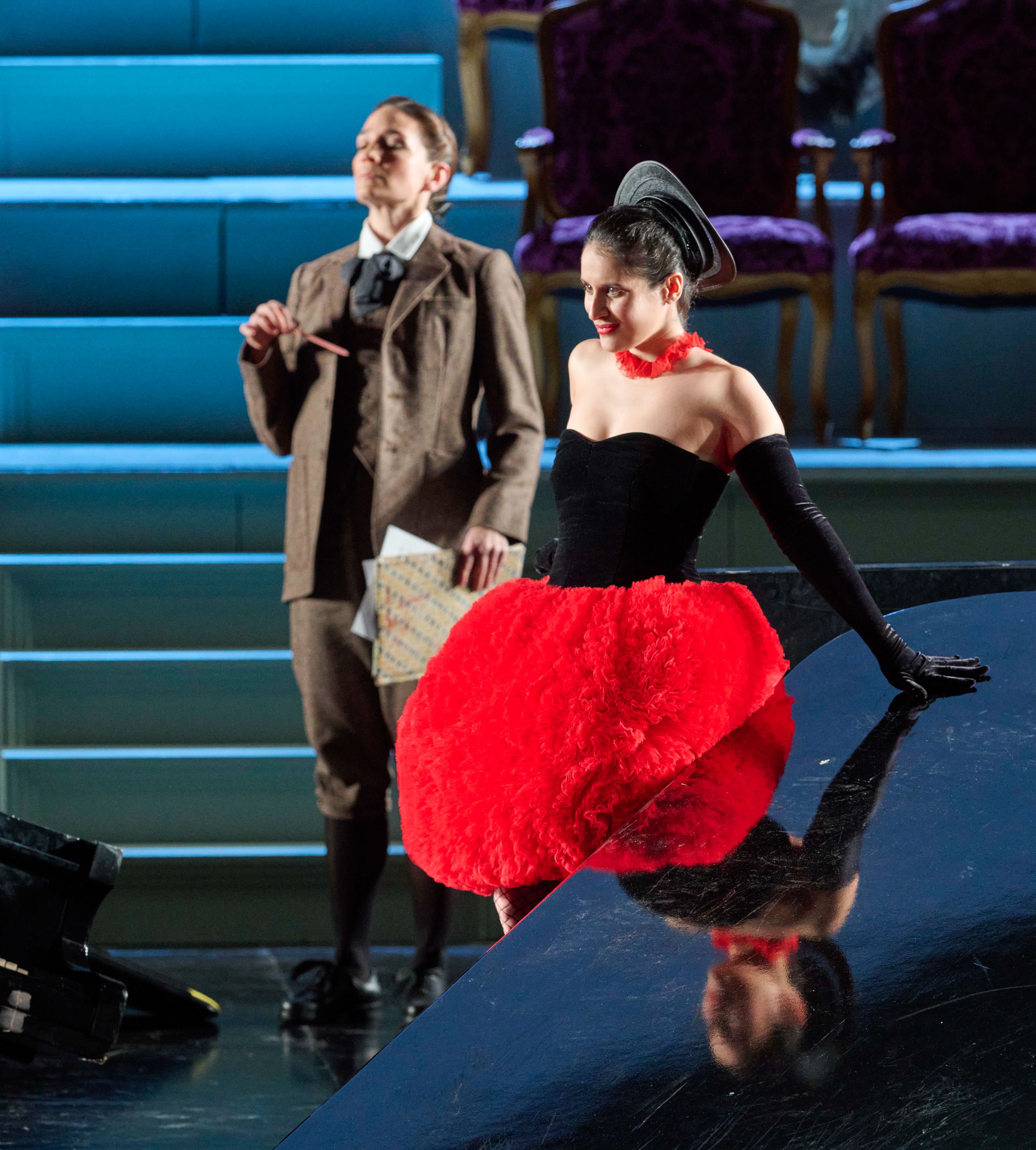 Eine Frau in schwarzem Korsett und rotem Tutu lehnt sich an einen Flügel, während eine Person in braunem Anzug daneben steht und eine Schriftrolle hält.