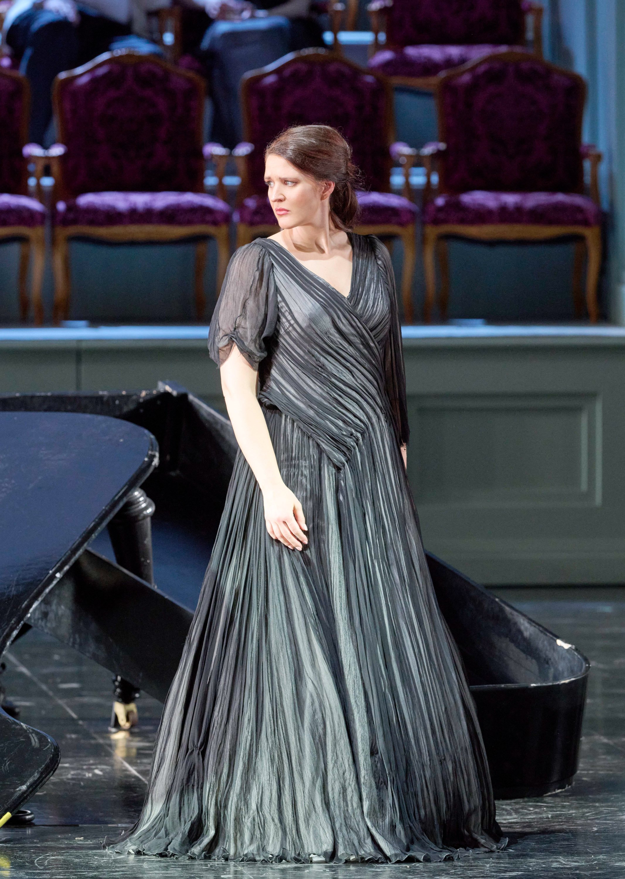 Eine Frau in einem eleganten, silbergrauen Kleid steht auf der Bühne neben einem schwarzen Flügel und blickt ernst zur Seite; im Hintergrund leere Theatersessel.