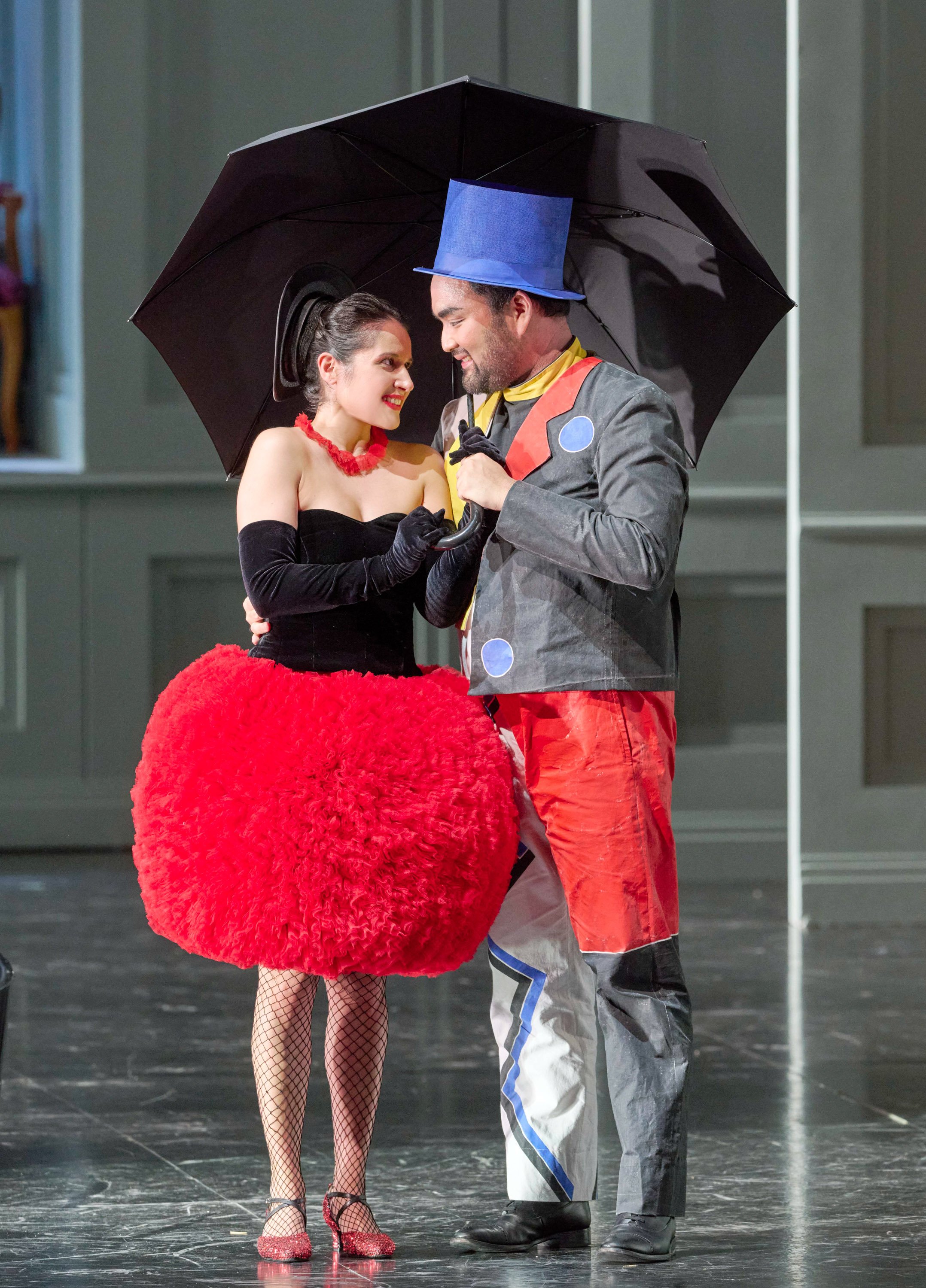 Eine Frau in rotem Tutu und ein Mann in bunter Verkleidung stehen eng beieinander unter einem schwarzen Regenschirm und blicken sich lächelnd an.