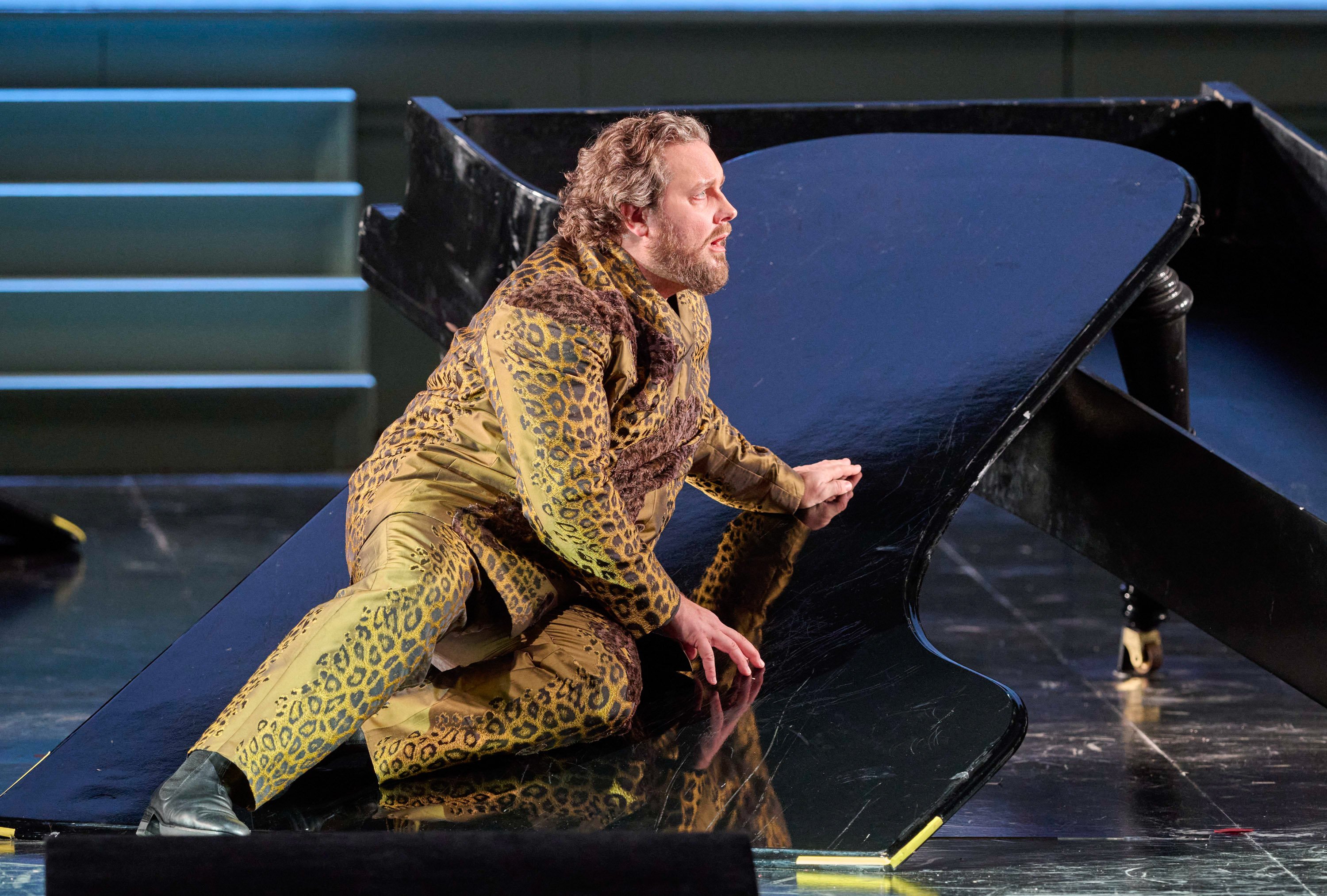 Ein Mann in golden gemustertem Anzug kniet auf einer Bühne neben einem schwarzen Flügel und schaut mit angespanntem Gesichtsausdruck zur Seite.