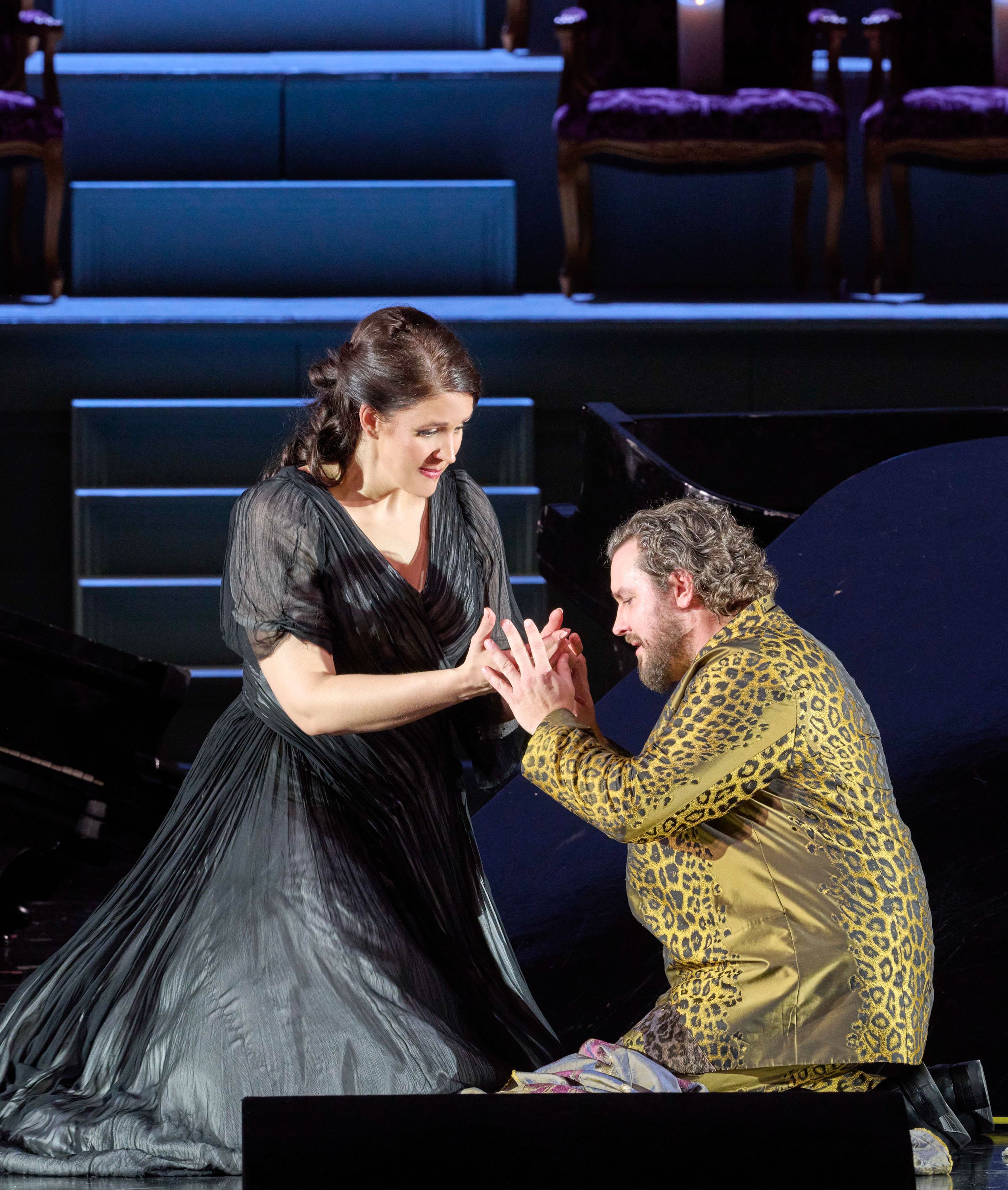 Eine Frau in einem eleganten dunklen Kleid und ein Mann in goldenem Anzug knien sich gegenüber und halten sich innig die Hände auf der Bühne.