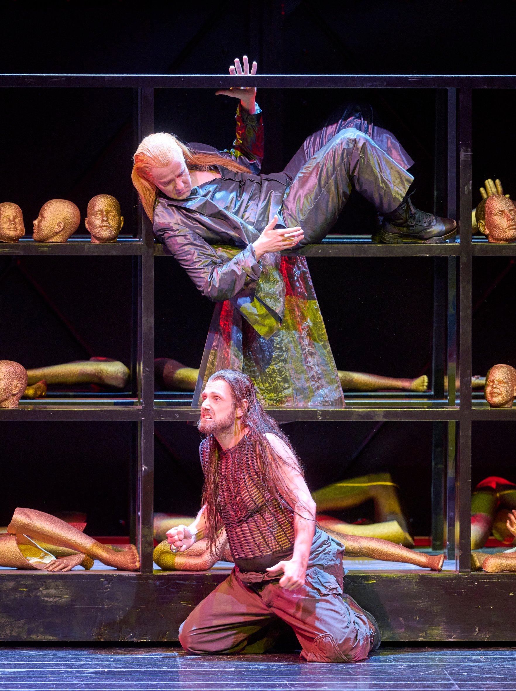 Eine Person in schillerndem Mantel liegt dramatisch in einem Regal voller Köpfe und Gliedmaßen, während eine weitere Figur unten kniet.