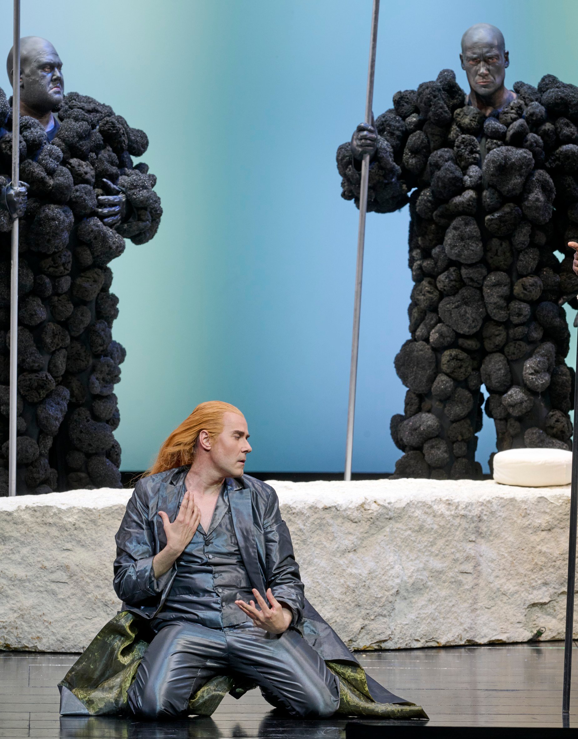 Eine Figur mit langem roten Haar kniet in silbernem Kostüm auf der Bühne und blickt dramatisch zur Seite; im Hintergrund stehen zwei schwarze, skulpturartige Gestalten.