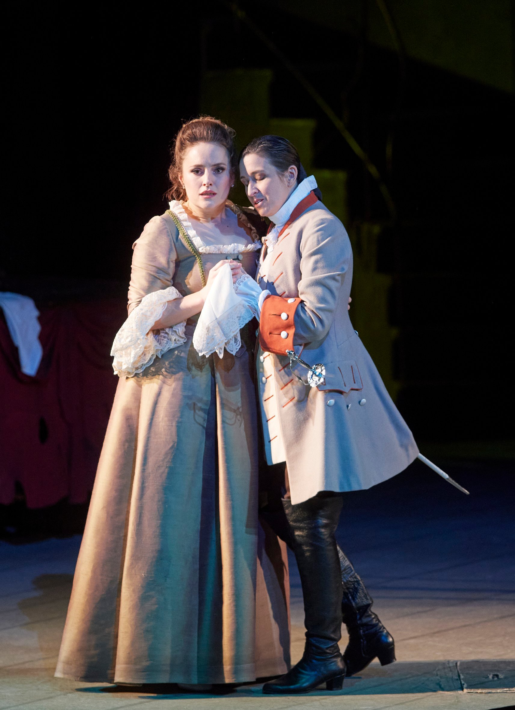 Eine Frau in einem braunen Kleid wird zärtlich von einer Person in historischer Uniform gehalten, beide blicken ernst in die Ferne.