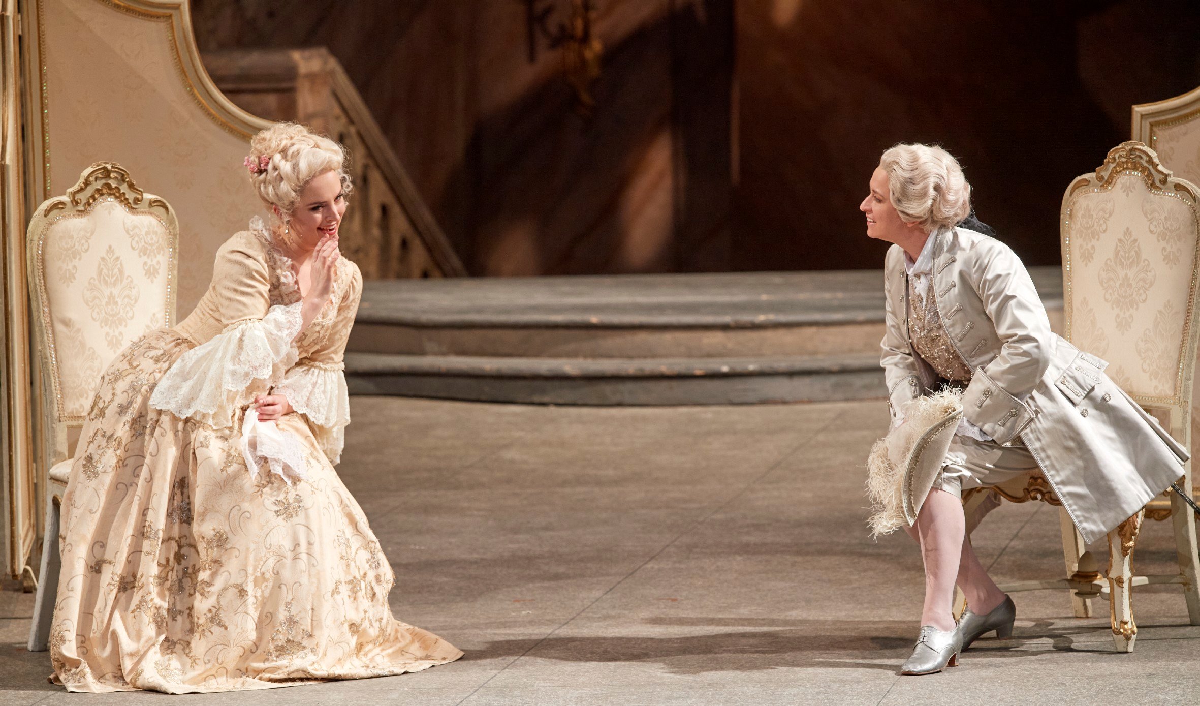 Eine Frau in einem cremefarbenen Rokoko-Kleid sitzt schmunzelnd, während eine andere Person sich in weißer historischer Kleidung zu ihr vorbeugt.