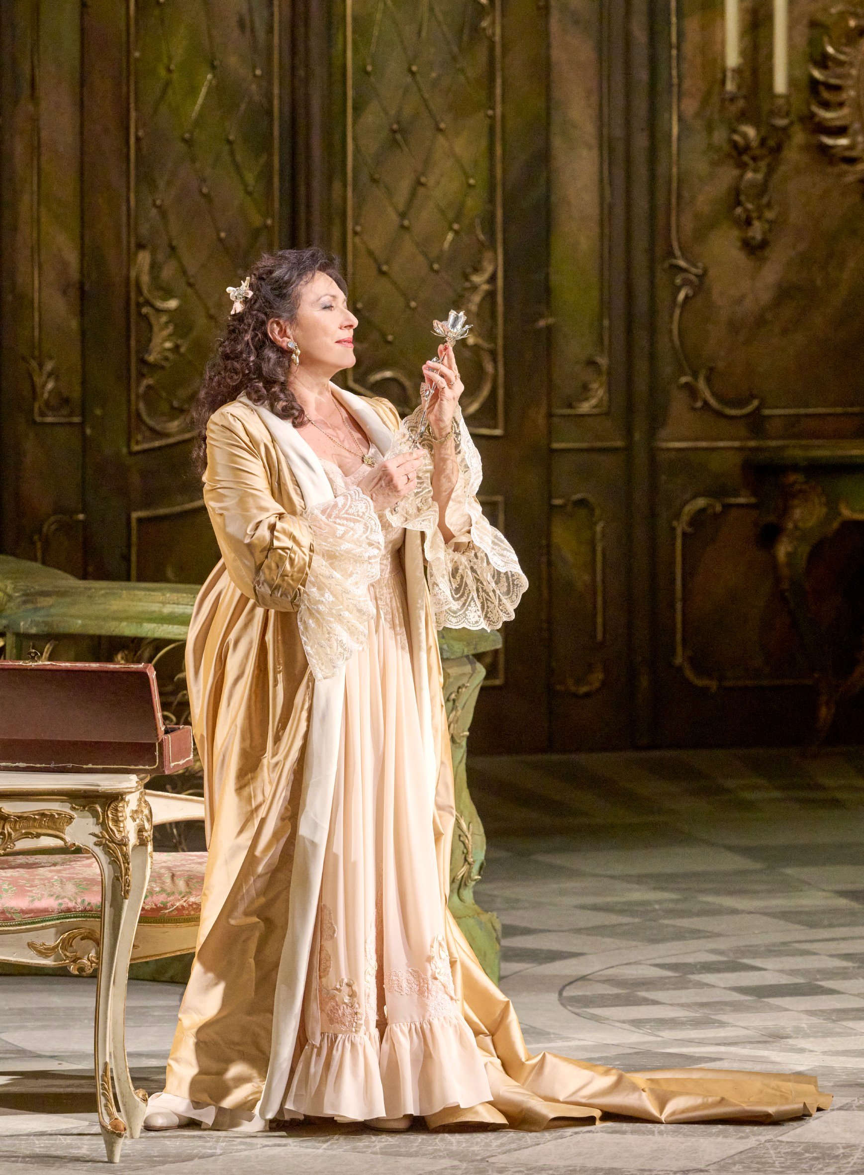 Eine Frau in einem eleganten, goldfarbenen Kleid steht vor einem Schminktisch und betrachtet nachdenklich ein kleines Kreuz.