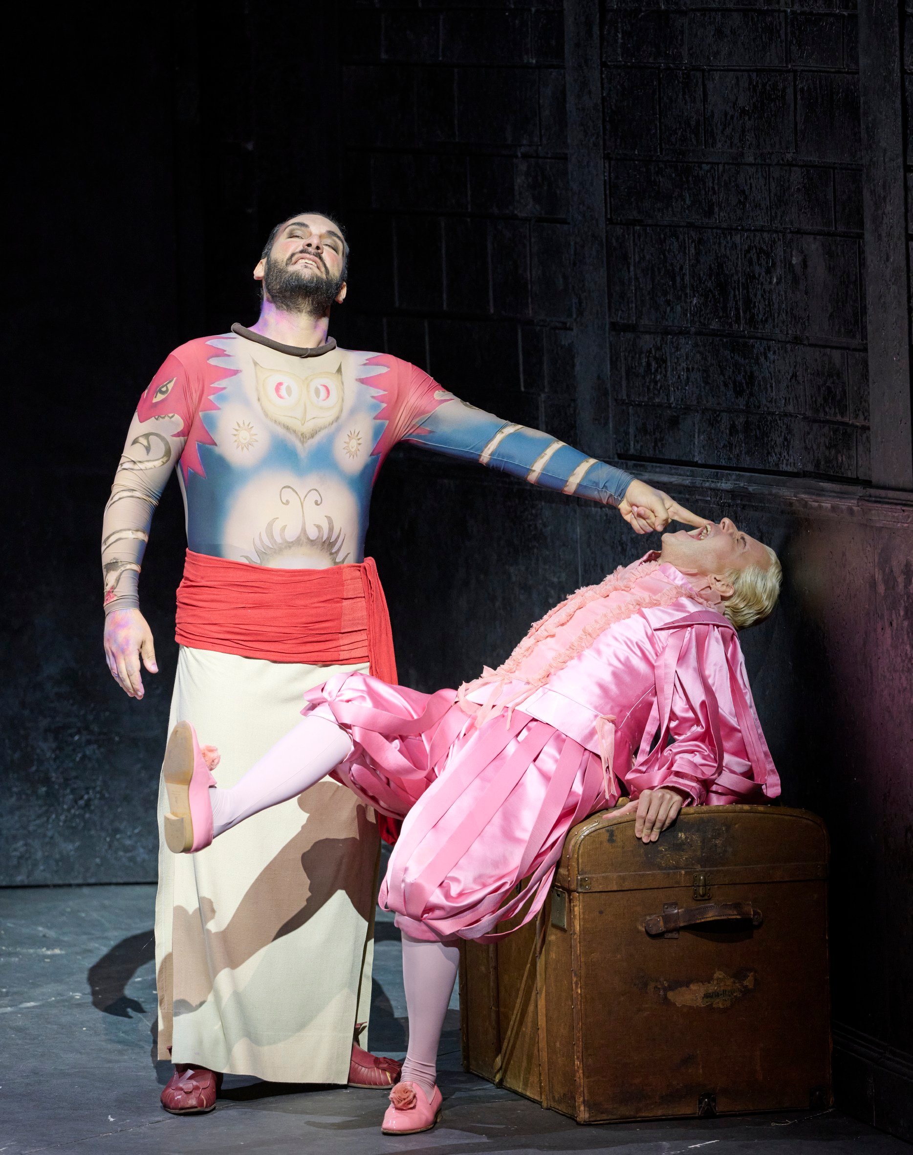 Ein tätowierter Mann mit roter Schärpe hält dramatisch das Kinn einer Person in pinkem Kostüm, die rücklings über einer Truhe liegt.