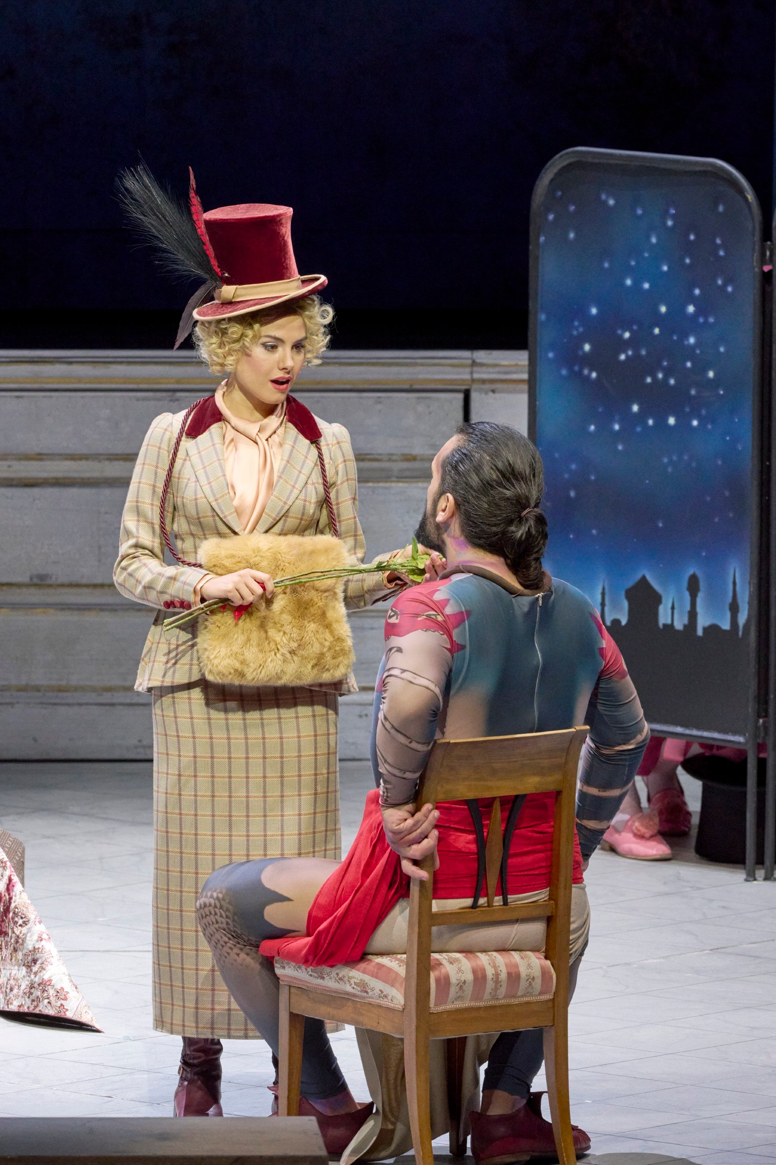 Eine Frau in gestreiftem Kostüm und rotem Zylinder steht vor einem Mann, der auf einem Stuhl sitzt.