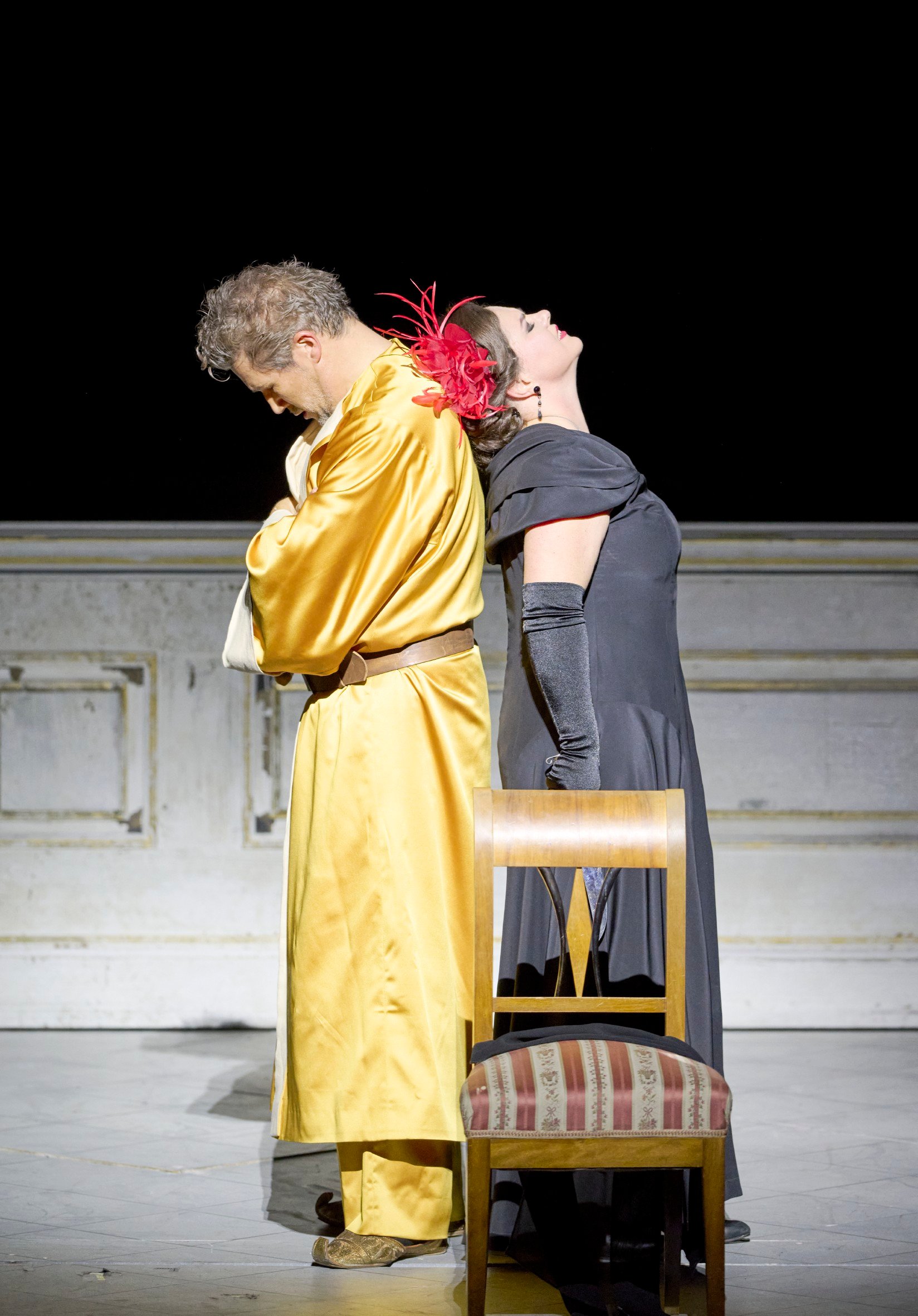 Ein Mann in gelbem Gewand und eine Frau in schwarzem Kleid mit rotem Federschmuck stehen Rücken an Rücken, voneinander abgewandt.