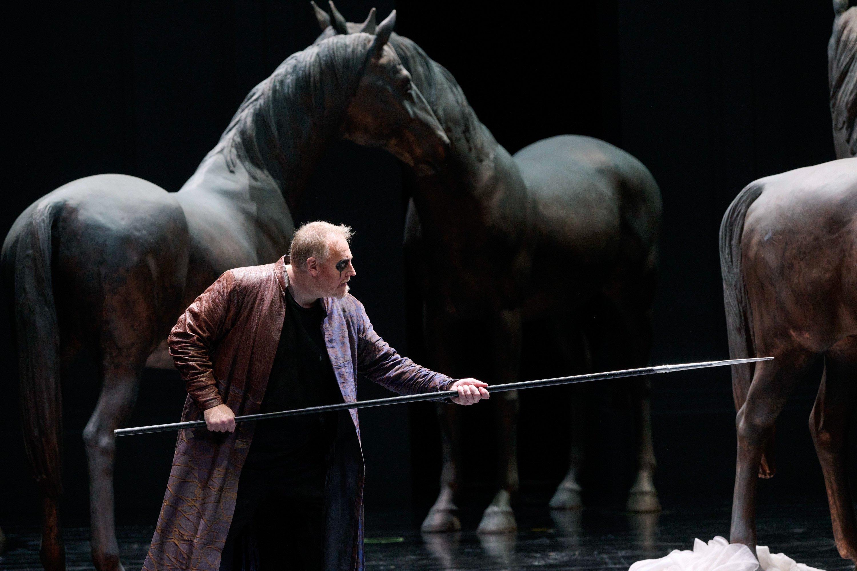 Ein Mann in langem Mantel hält einen langen Stab, während im Hintergrund große Pferdefiguren stehen.