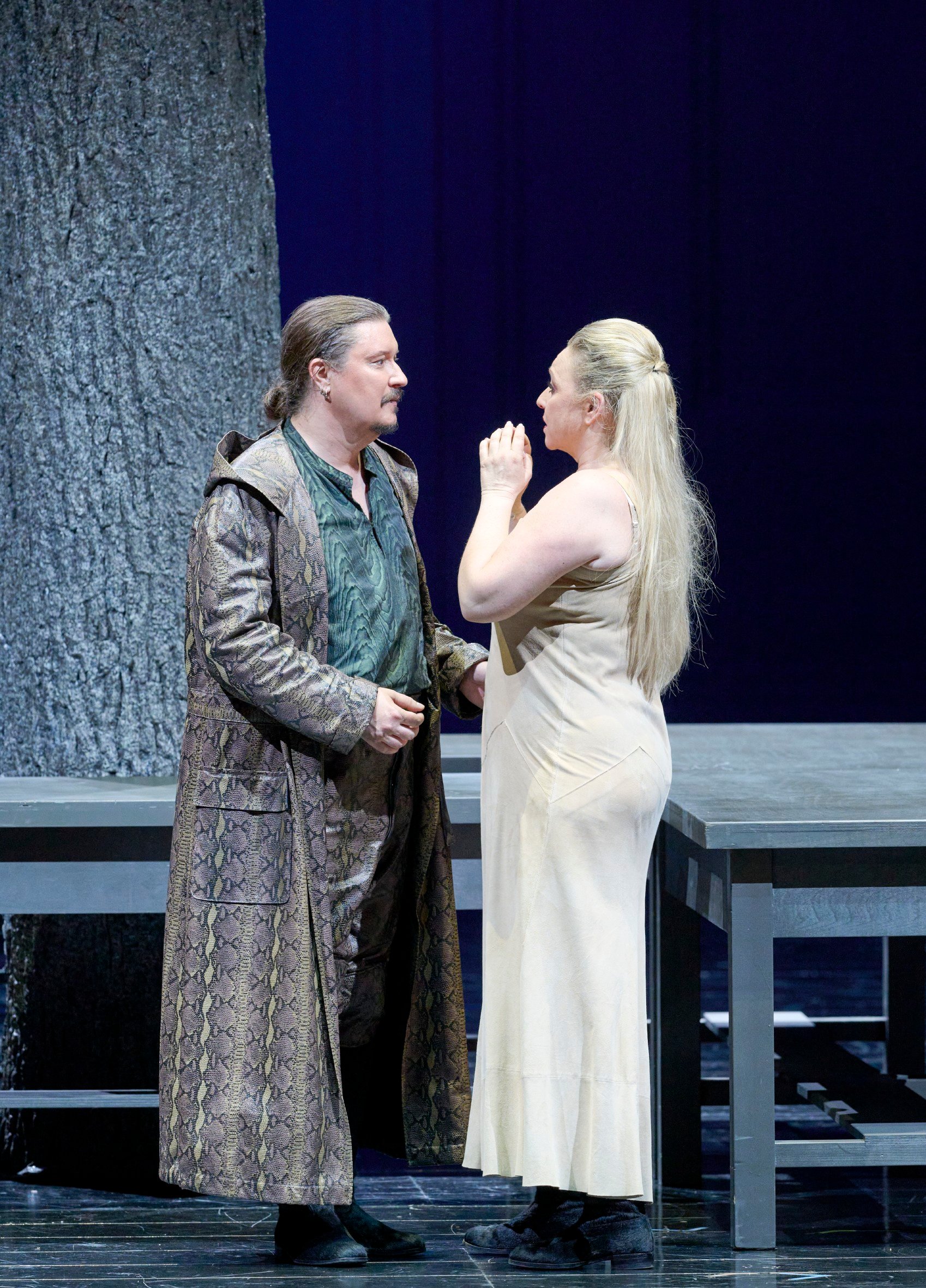 Ein Mann in langem Mantel steht einer Frau im beigefarbenen Kleid gegenüber, beide wirken emotional bewegt.