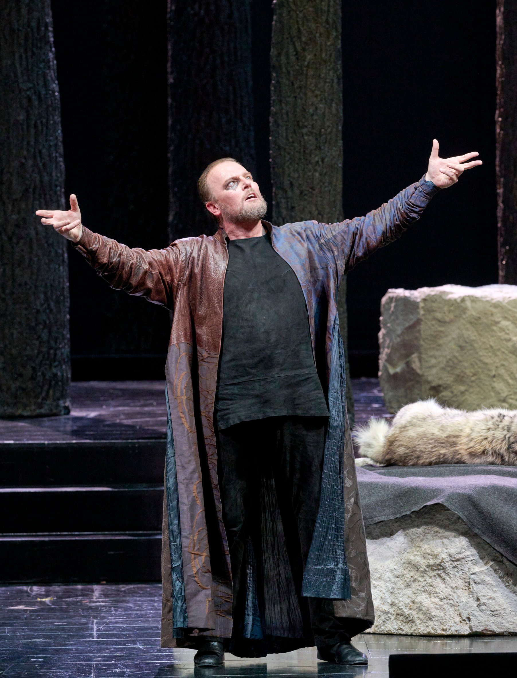 Ein Mann in langem Mantel steht mit ausgebreiteten Armen auf der Bühne, umgeben von Felsen und Baumstämmen.
