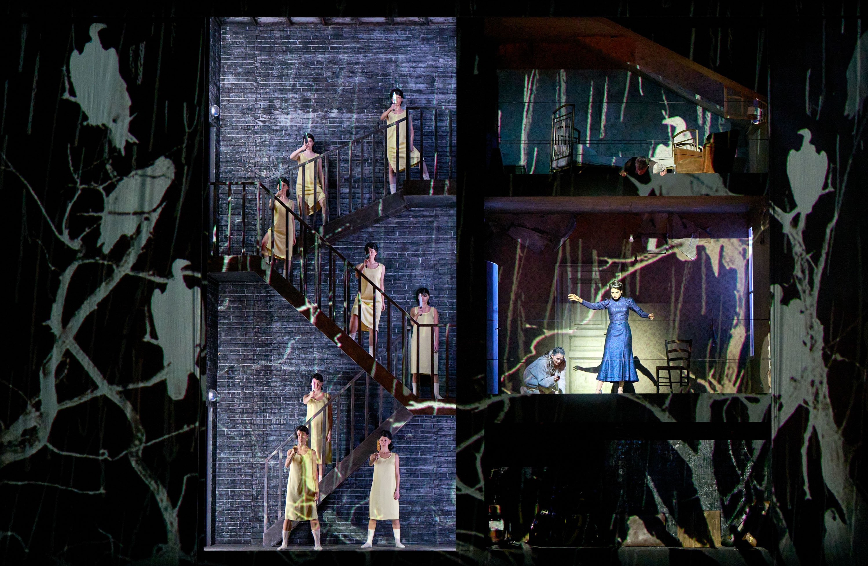 Auf einer Bühne sind mehrere Stockwerke eines Hauses abgebildet, mit Darstellern in weißen Kleidern, die in verschiedenen Posen auf Treppen und in Räumen stehen.