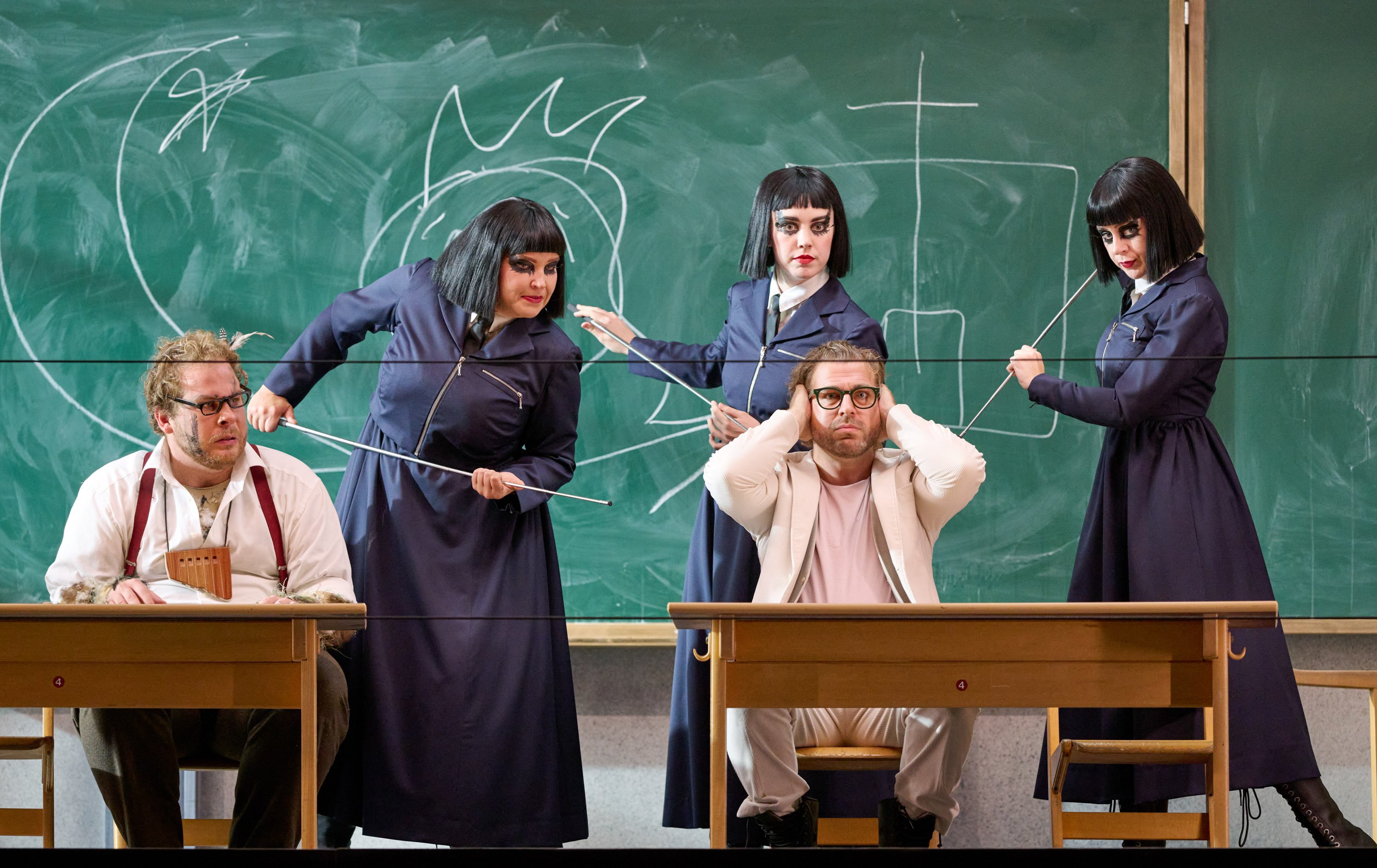 Drei Frauen mit schwarzen Haaren langen dunkelblauen Gewändern stehen in einem Klassenzimmer. Vor ihnen sitzen zwei Männer in hellen Gewändern an Tischen. Einer von ihnen hält sich die Ohren zu.