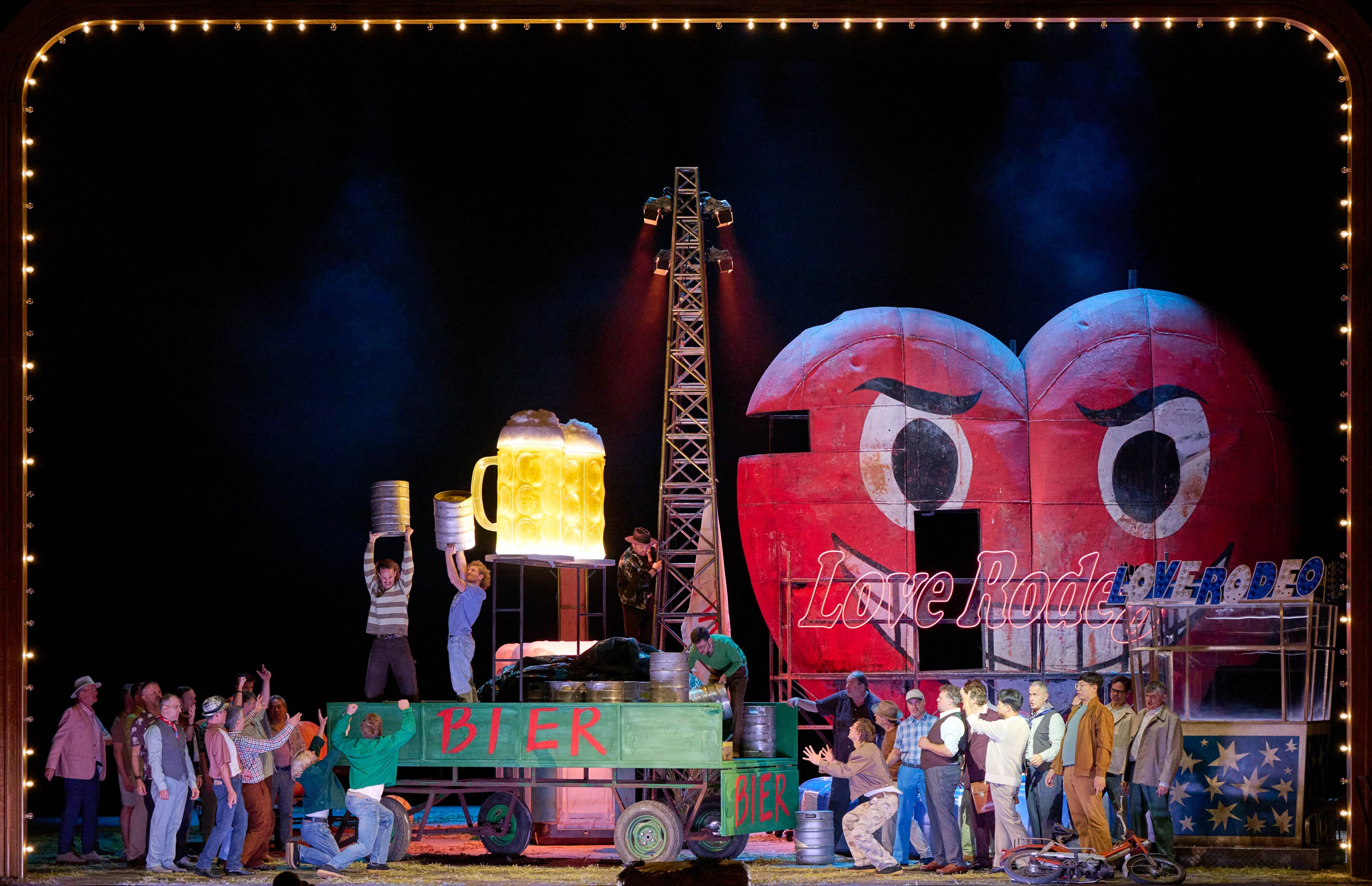 Auf einem Anhänger mit der Aufschrift "Bier" stehen zwei Männer mit riesigen Biergläsern. Neben dem Wagen stehen viele Menschen, die zu den beiden Männern hinaufblicken. Im Hintergrund sieht man die Stände einer Kirmes.