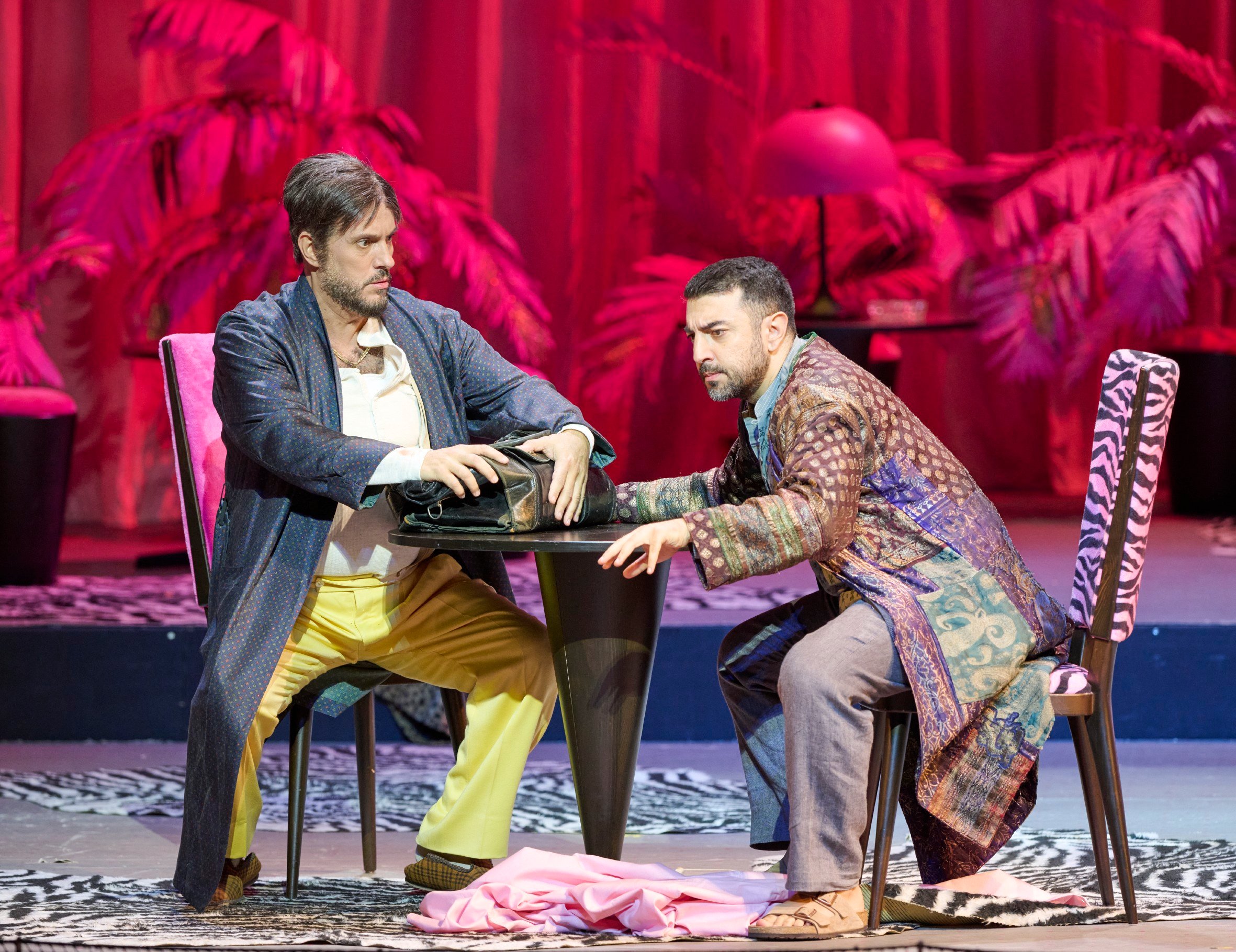 Zwei Männer sitzen an einem runden Tisch vor rotem Hintergrund.