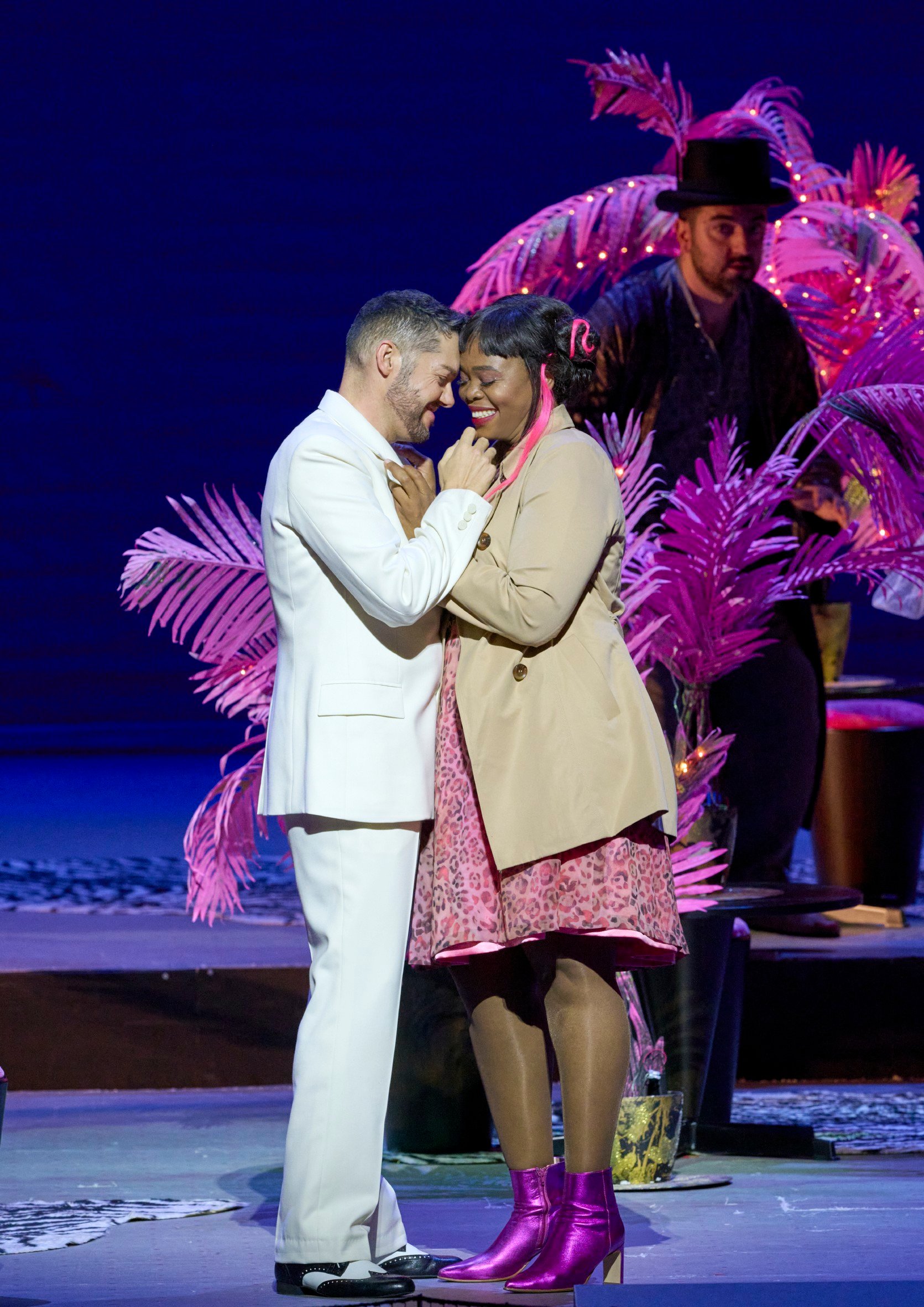 Ein Mann im weißen Anzug steht Stirn an Stirn mit einer Frau; im Hintergrund eine bunte Szenerie.