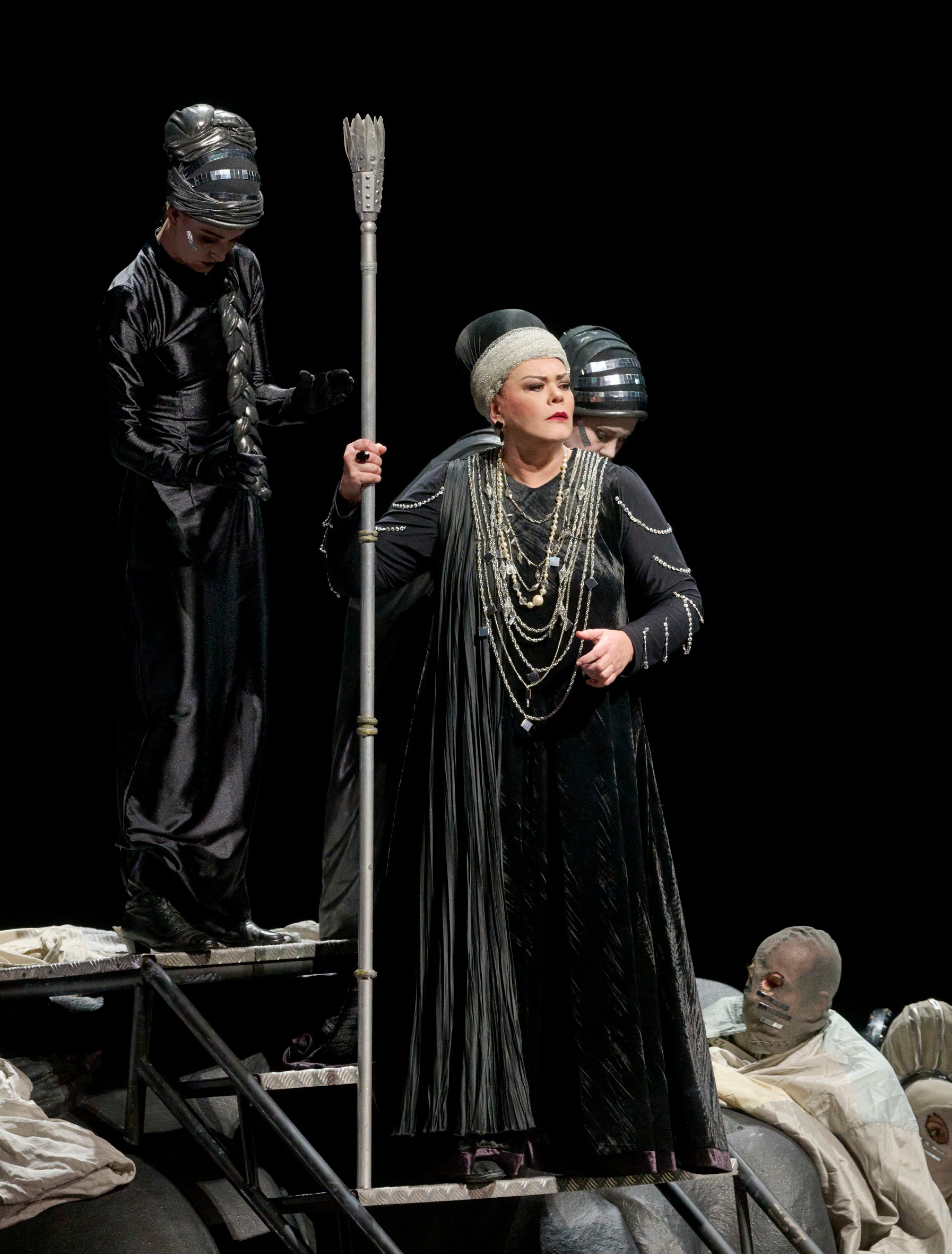 Eine zentrale Figur mit Stab und Perlenschmuck steht auf der Bühne, begleitet von zwei dunkel gekleideten Personen mit glänzenden Kopfbedeckungen.