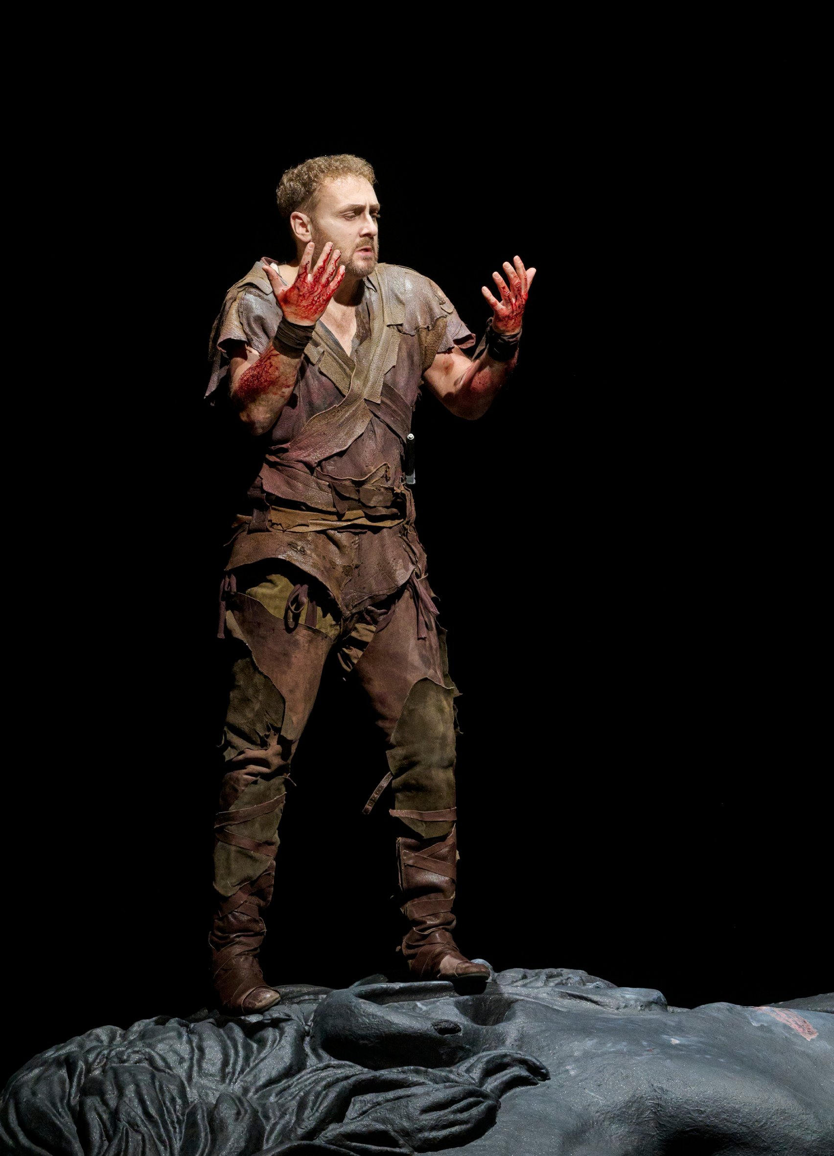 Ein Mann im zerlumpten Kostüm steht mit blutverschmierten Händen dramatisch auf der Bühne.