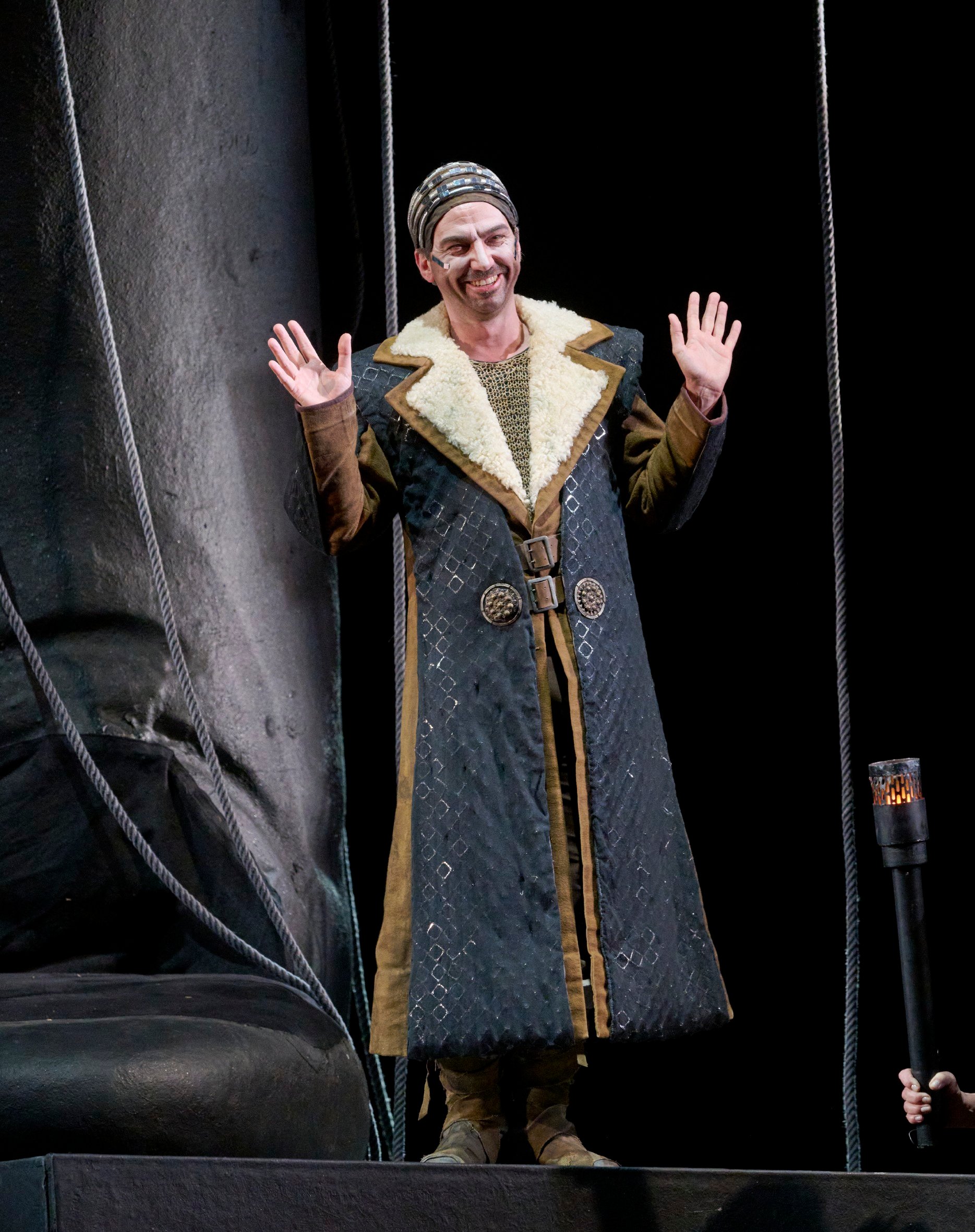 Ein Mann in einem langen, reich verzierten Mantel steht lächelnd auf der Bühne und hebt beide Hände.