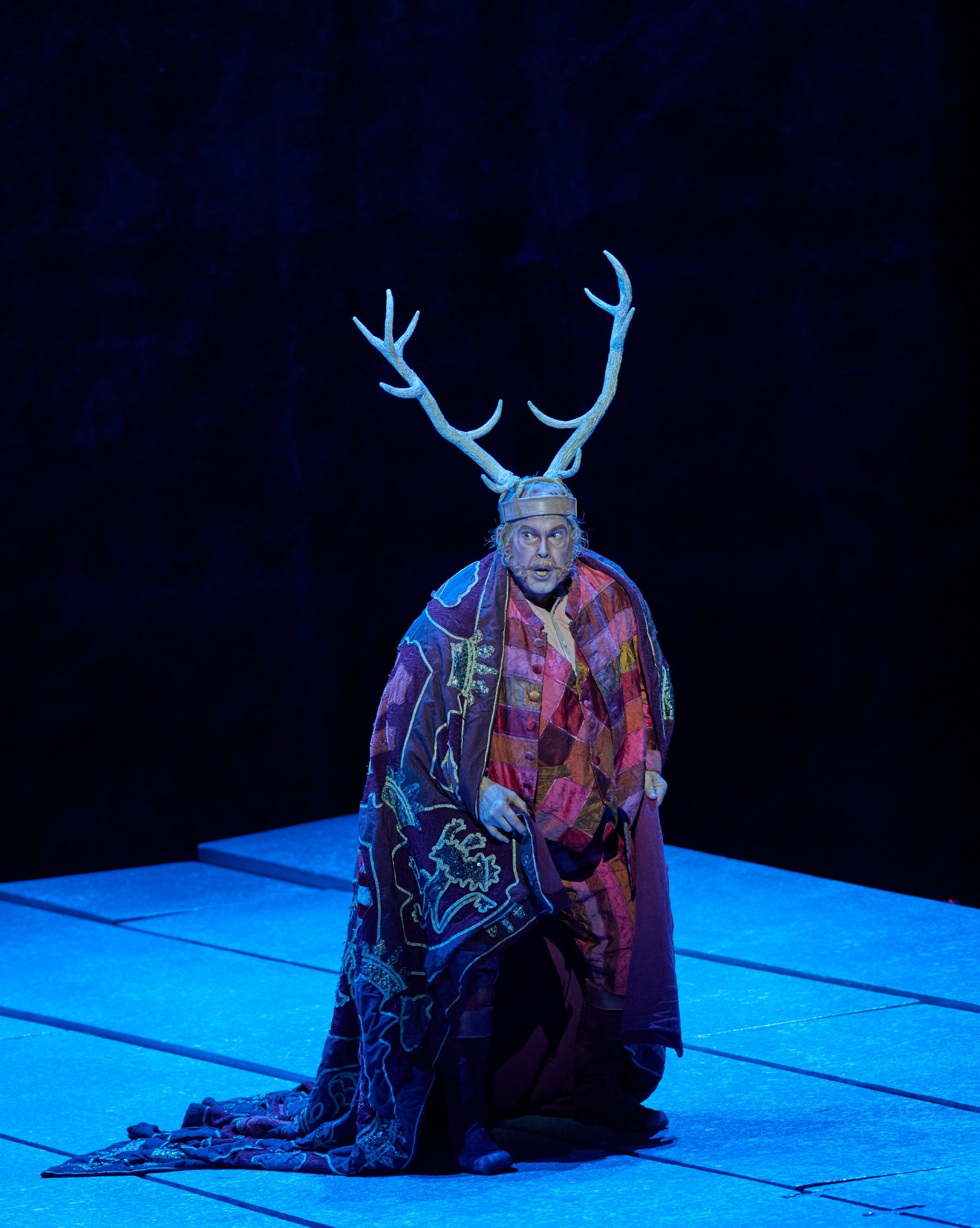Eine Person mit Geweih und farbenfrohem Umhang steht auf einer blau beleuchteten Bühne vor dunklem Hintergrund.