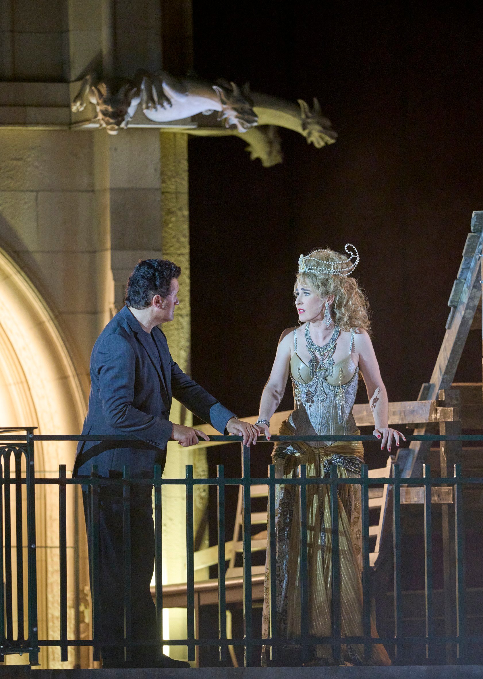 Ein Mann und eine Frau stehen sich auf einem Balkon gegenüber; er spricht sie an, sie hört aufmerksam zu, umgeben von theatralischer Kulisse.