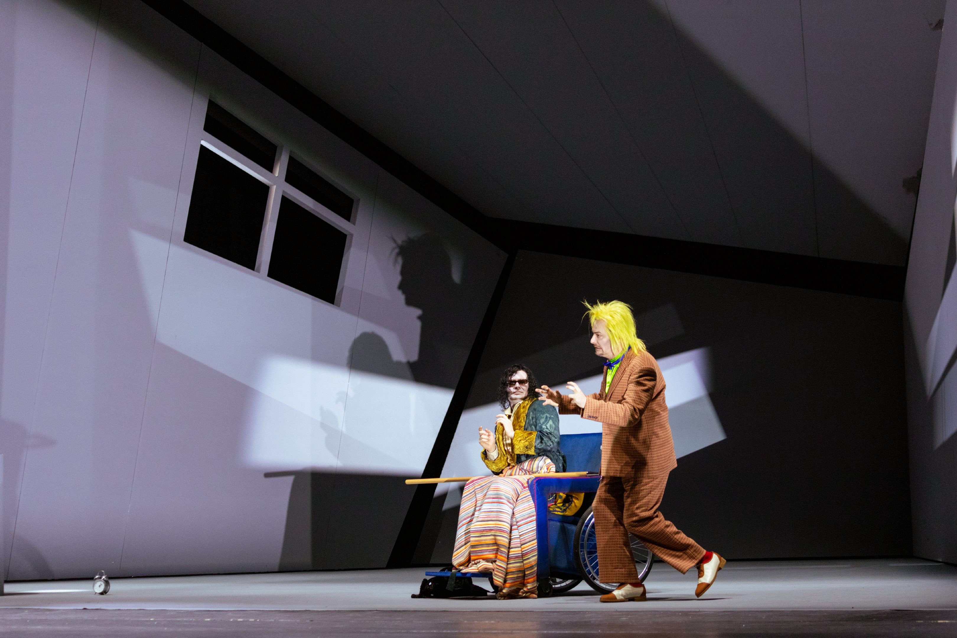 Eine bunt gekleidete Person sitzt im Rollstuhl, während eine weitere Figur im Tierkostüm lebhaft davor agiert, begleitet von Schattenprojektionen an der Wand.