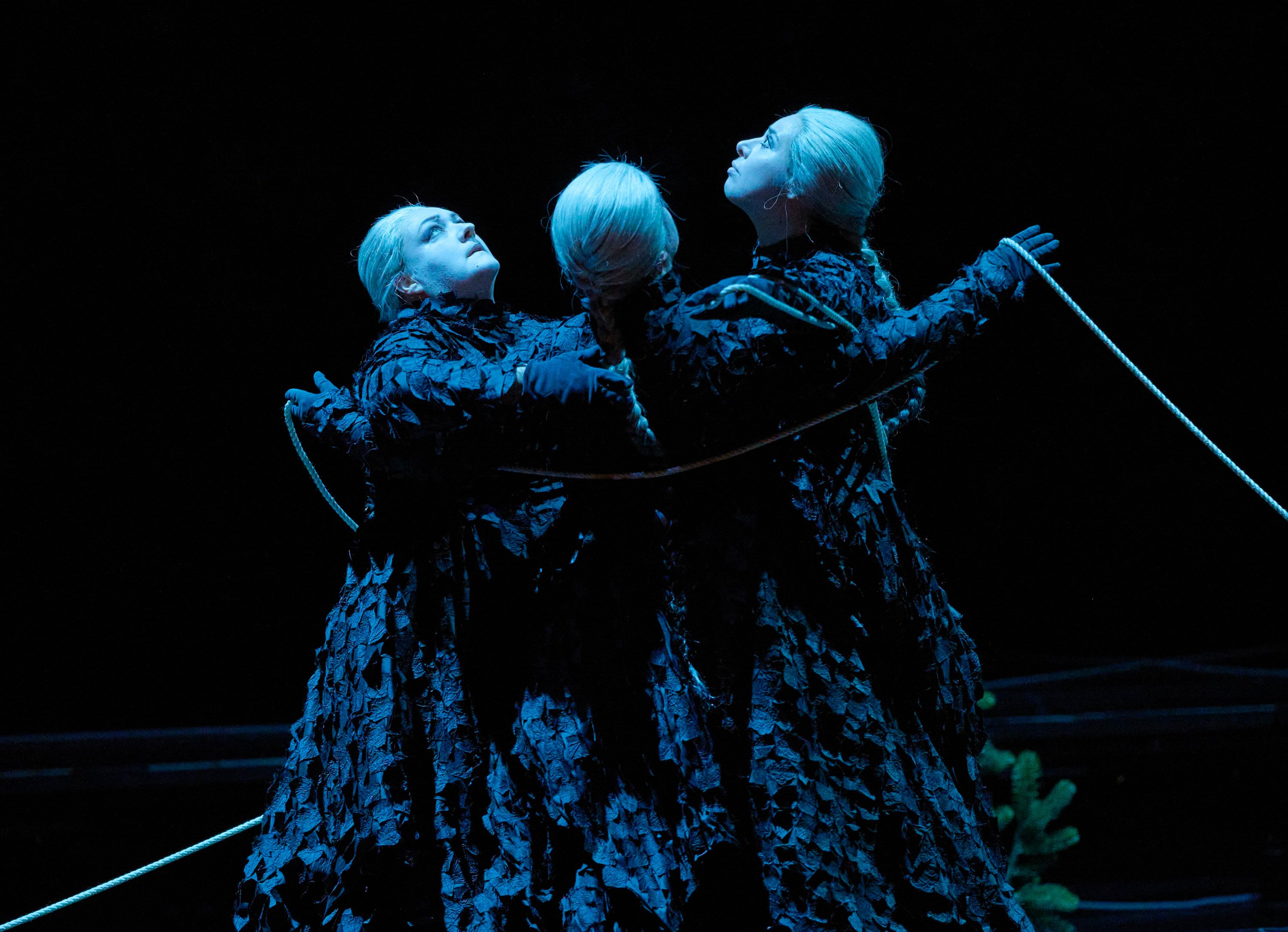 Drei weißhaarige Frauen in dunklen Gewändern stehen eng umschlungen und halten ein Seil.