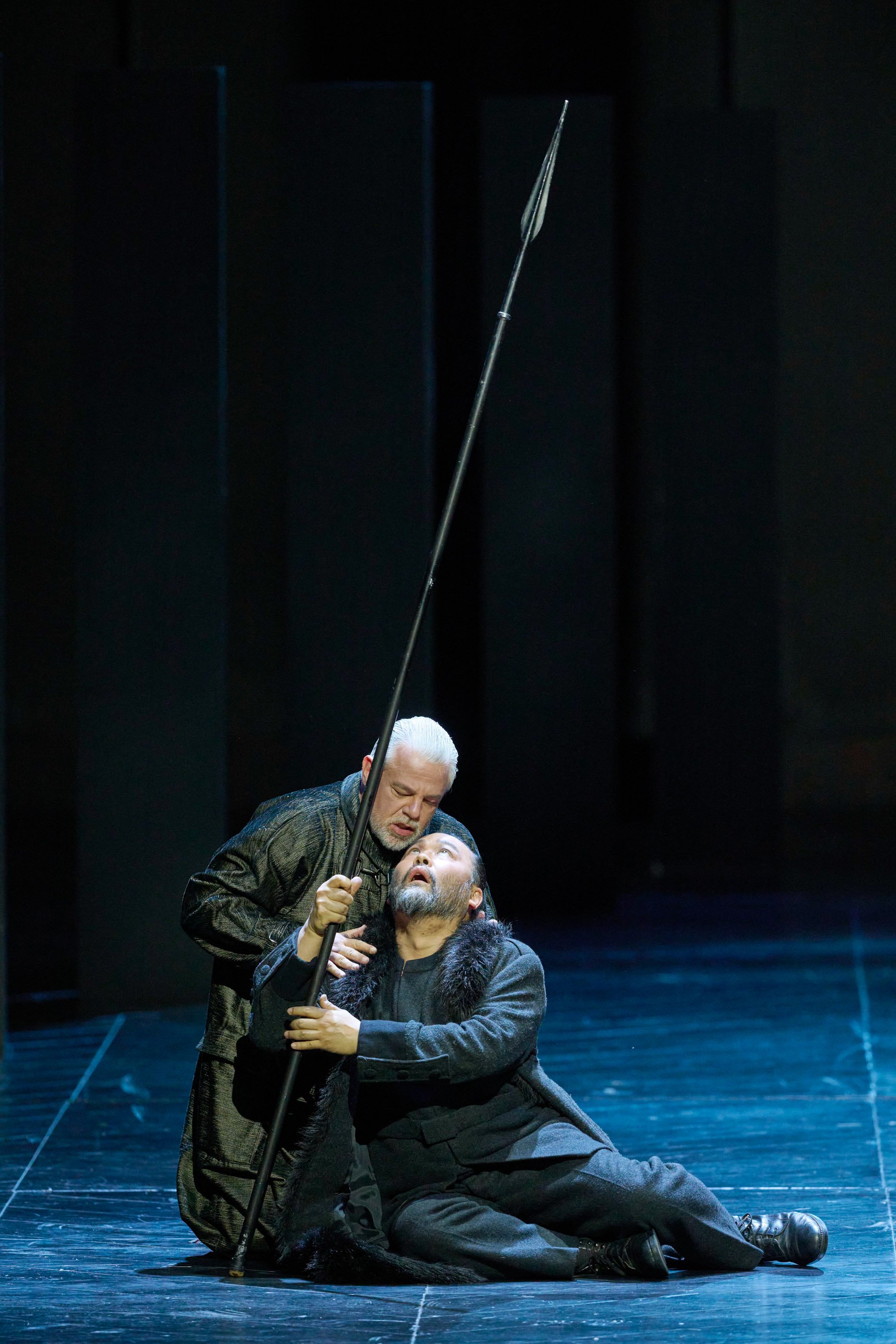 Ein Mann liegt auf dem Boden während er eine Lanze in die Höhe hält; er wird von einem anderen Mann an der Schulter gehalten.