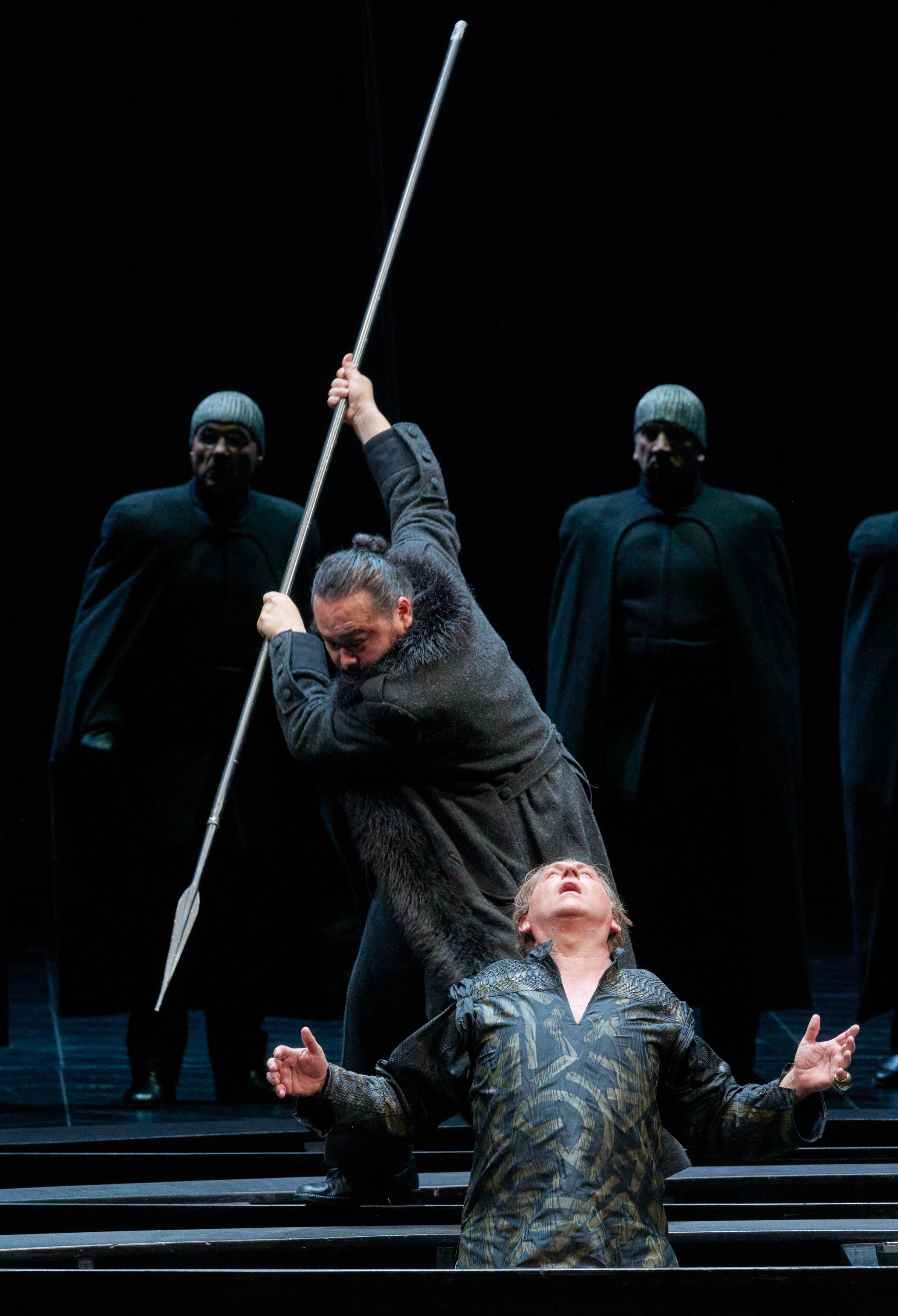 Ein Mann hebt einen Speer bedrohlich über einem anderen, während schwarz gekleidete Gestalten im Hintergrund stehen.