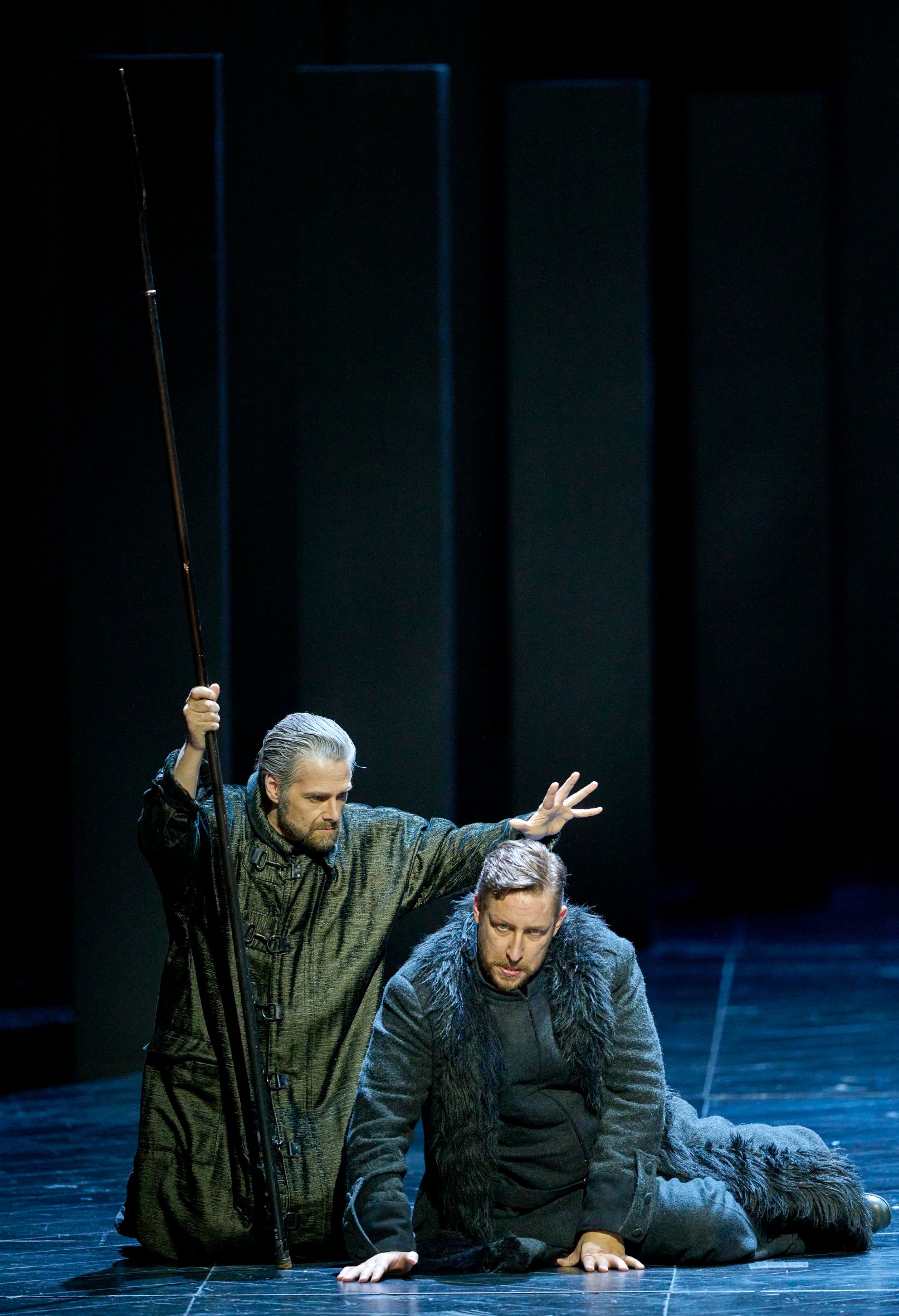 Zwei Männer in dunklen Gewändern befinden sich auf einer düsteren Bühne; einer kniet mit gesenktem Blick, während der andere eine segnende oder beschwörende Geste über ihn macht.