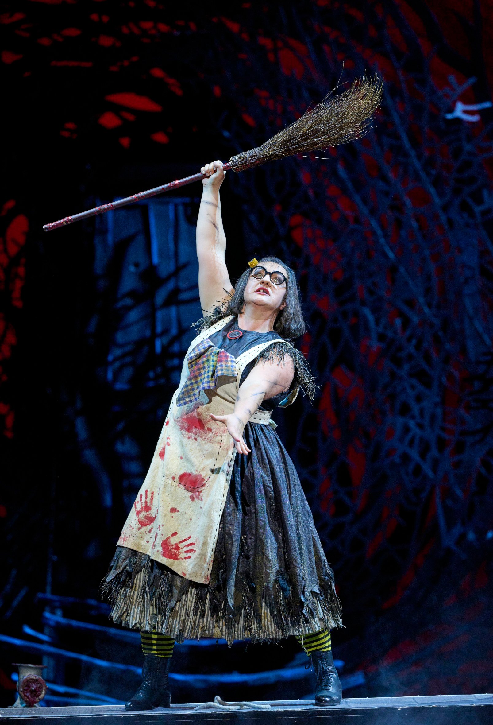 Eine theatralisch gekleidete Figur mit blutverschmierter Schürze hebt triumphierend einen Besen in die Luft vor düsterem Hintergrund.