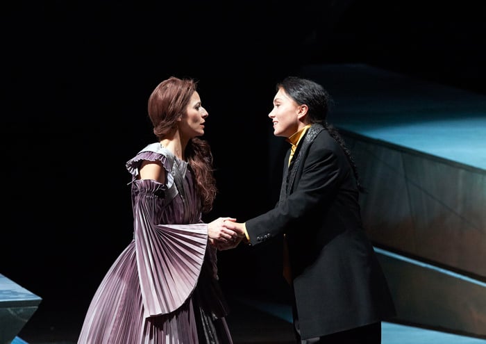 Zwei Personen stehen sich auf der Bühne gegenüber und halten sich an den Händen – die eine in einem historischen Kleid, die andere in modernerem Anzug.