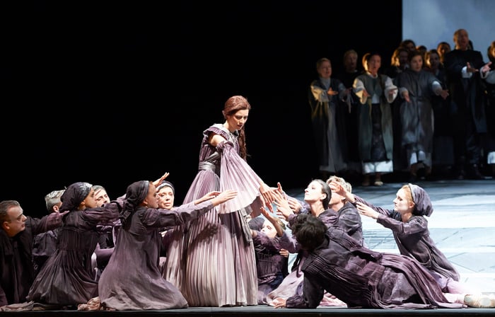 Eine Frau in einem historischen Kleid steht im Zentrum der Bühne, während zahlreiche Personen in ähnlicher Kleidung sie dramatisch umringen und nach ihr greifen.