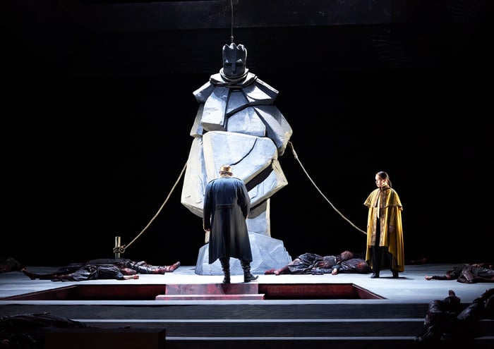 Eine imposante Statue steht im Zentrum der Bühne, umgeben von am Boden liegenden Figuren, während zwei Männer in dunklen Mänteln davor stehen.