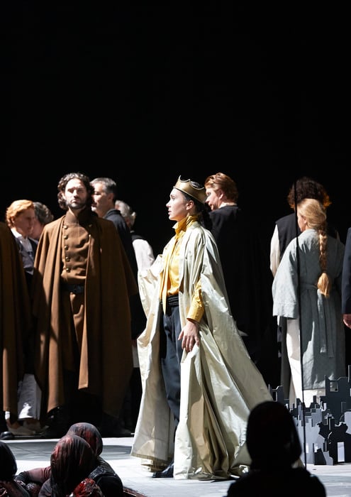 Eine Gruppe von Darstellern in historischen Gewändern steht auf der Bühne; eine zentrale Figur mit Krone und Umhang hebt den Blick würdevoll.