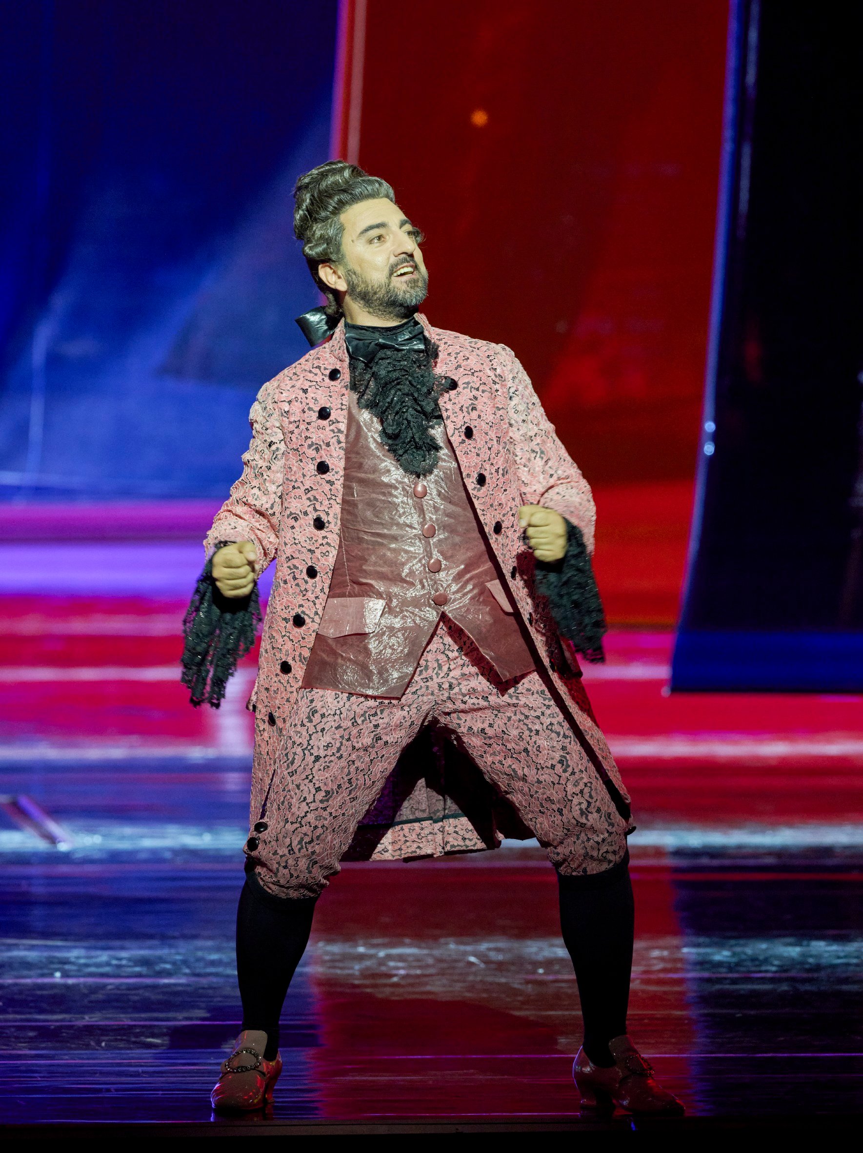 Ein Darsteller in auffälligem rosa Rokoko-Kostüm mit schwarzer Spitze steht auf der Bühne und singt mit leidenschaftlichem Ausdruck.