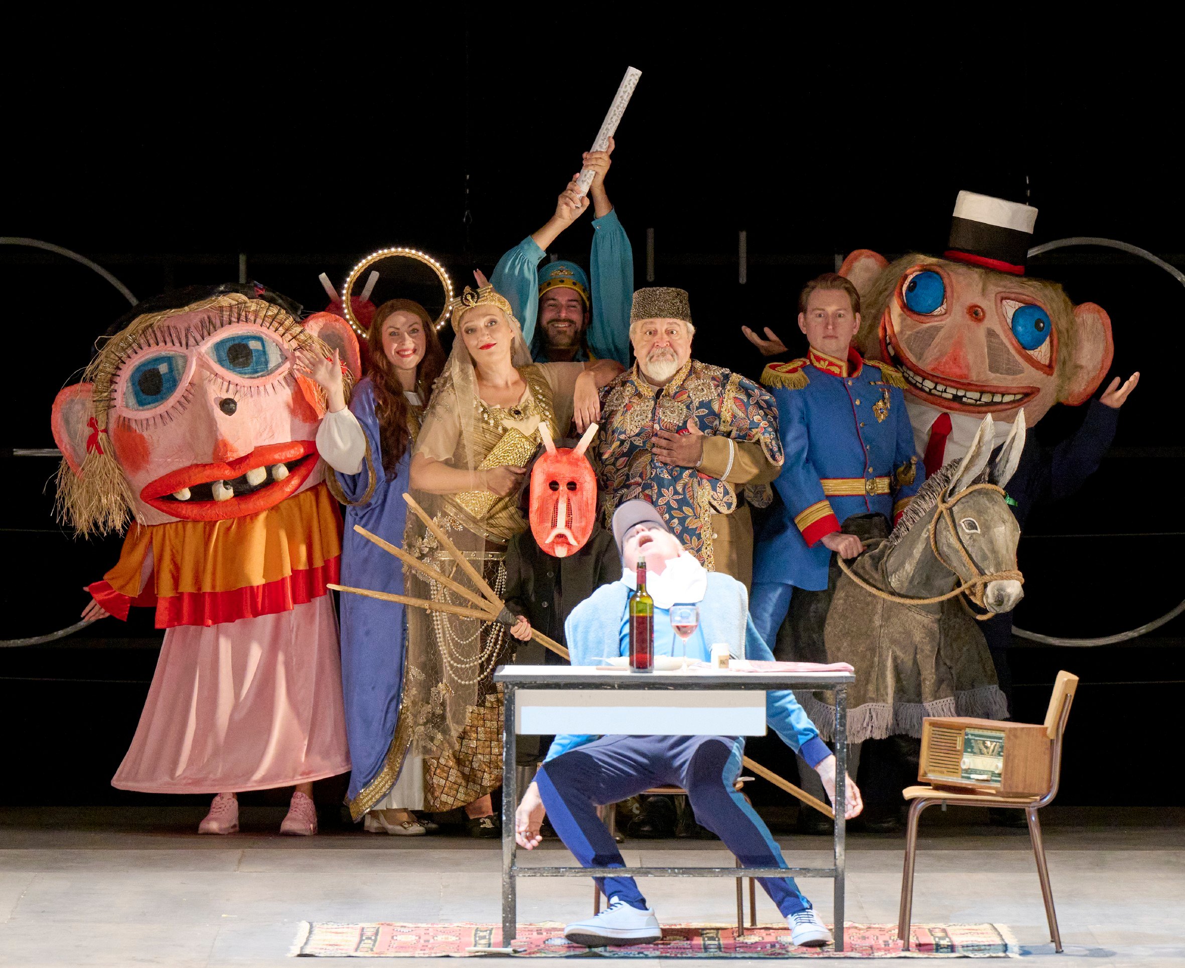 Eine bunte Gruppe von Darsteller:innen mit riesigen Masken, Kostümen und Requisiten posiert theatralisch um eine Figur, die leblos an einem Tisch sitzt.