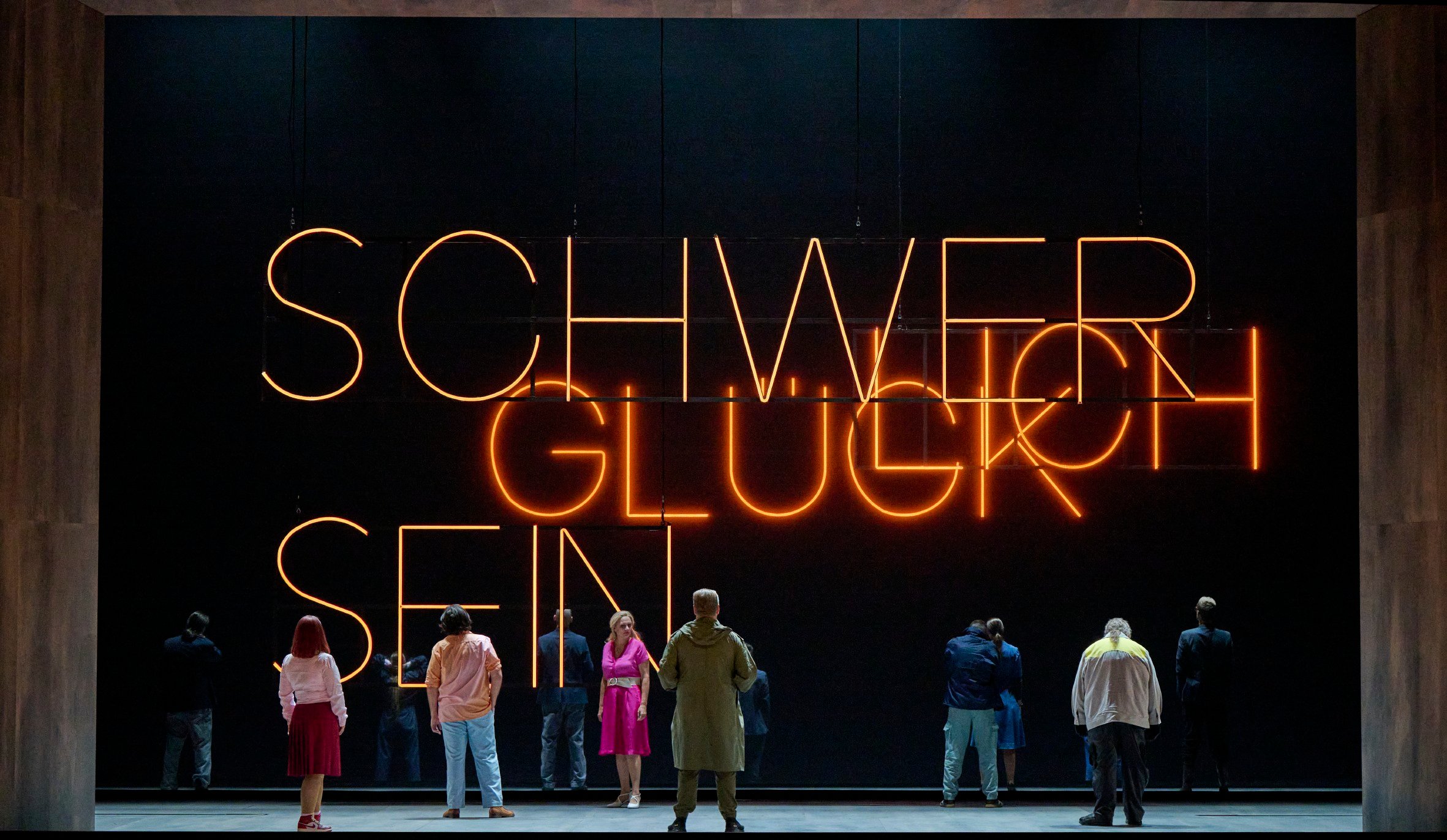 Mehrere Personen stehen auf einer Bühne vor einer riesigen Leuchtschrift mit den Worten „SCHWER GLÜCK SEIN“, teils nachdenklich, teils abgewandt.