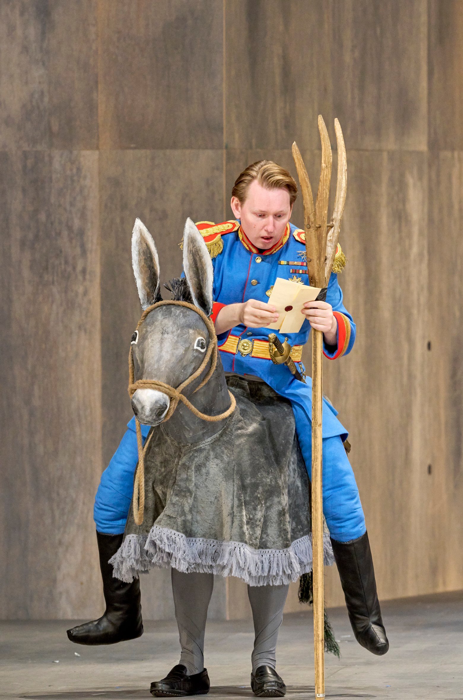 Ein Mann in blauer Uniform sitzt auf einem stilisierten Esel und liest überrascht einen Brief auf der Theaterbühne.